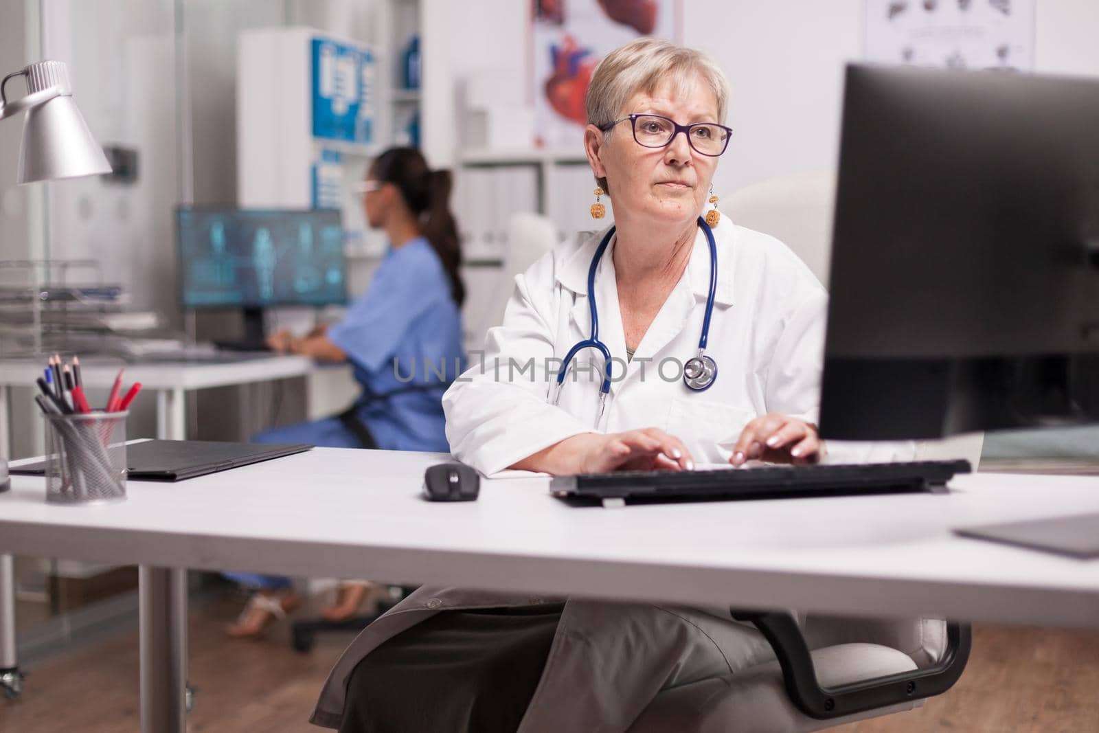 Experienced doctor using computer in clinic cabinet and nurse typing on pc in the background.