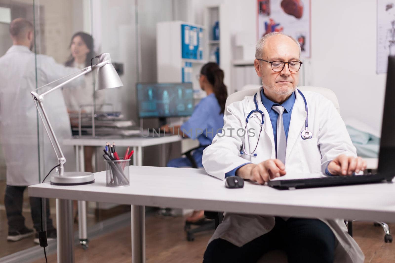 Doctor with grey hair in clinic cabinet working on computer and young medic discussing with woman patient on hospital corridor.