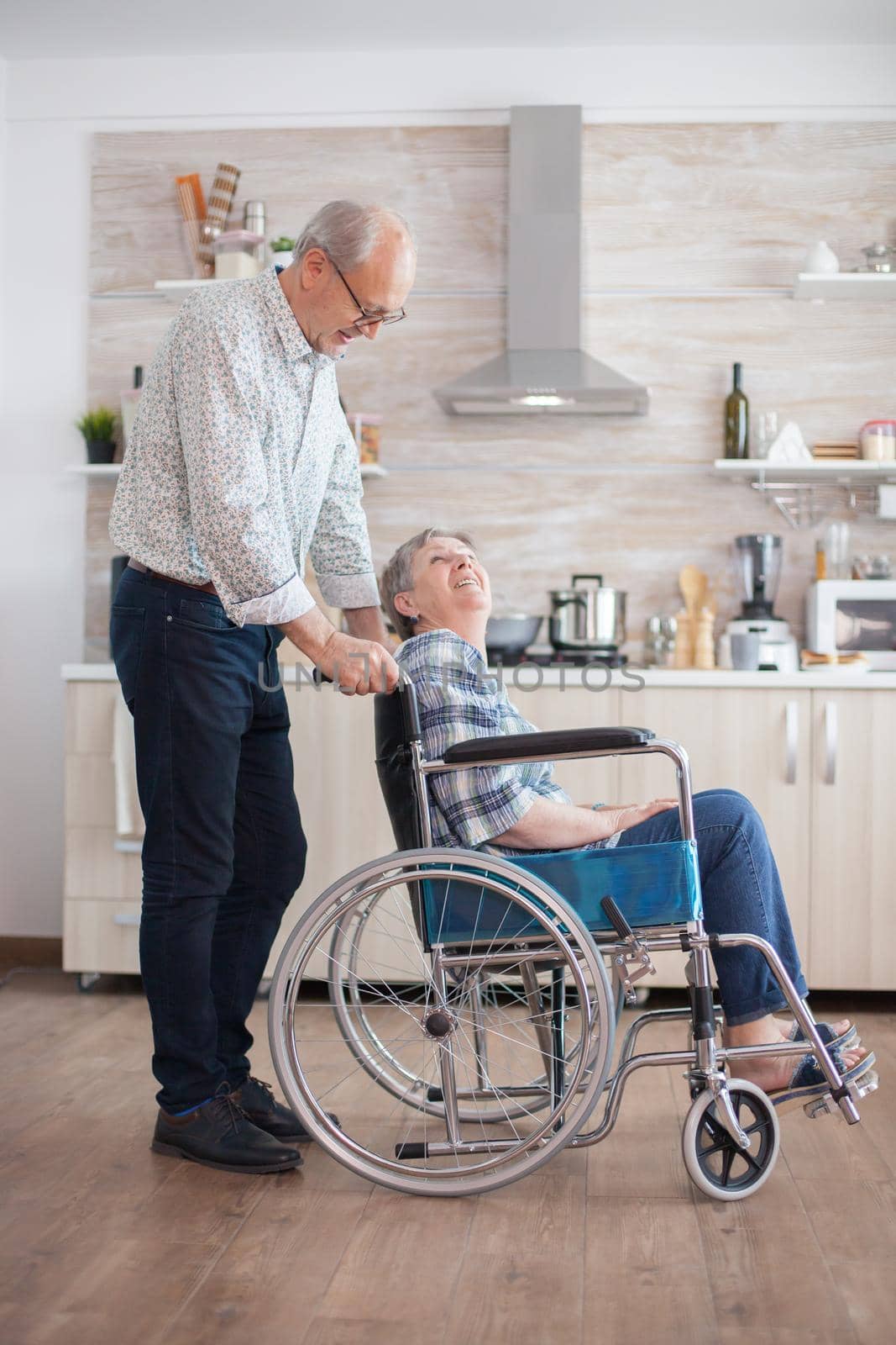 Husband looking at disabled senior woman in the kitchen. Disabled senior woman sitting in wheelchair in kitchen looking through window. Living with handicapped person. Husband helping wife with disability. Elderly couple with happy marriage.