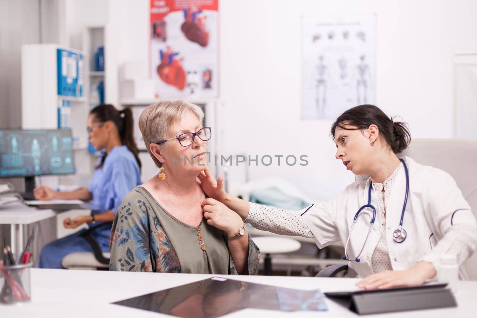 Doctor in hospital cabinet examining neck glands of senior patient. Mature woman with thyroid discomfort. Nurse in blue uniform working on computer.