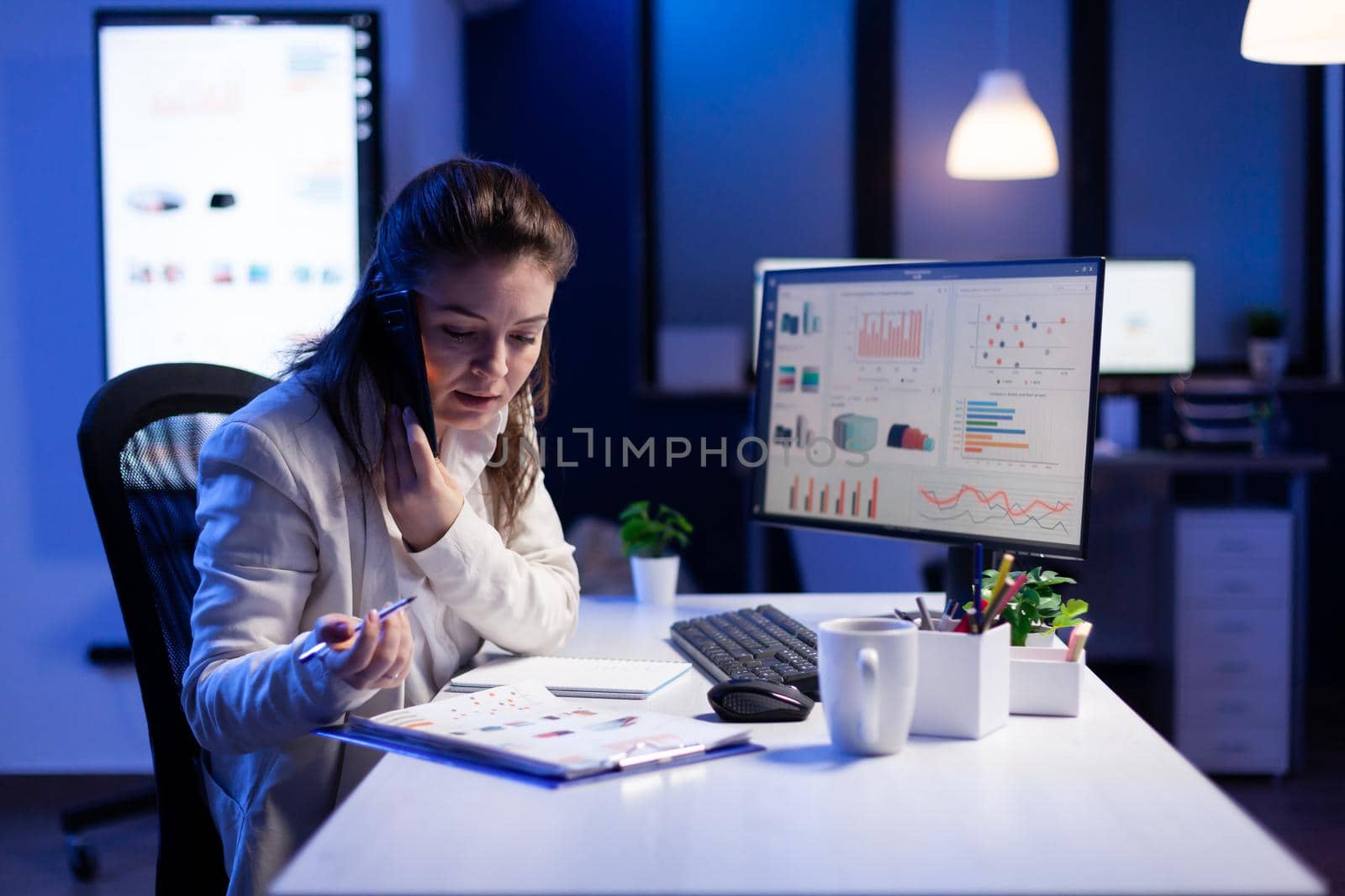 Businesswoman checking financial graphs while speaking with her team on smartphone late at night in company office. Busy manager using modern technology network wireless searching business solution