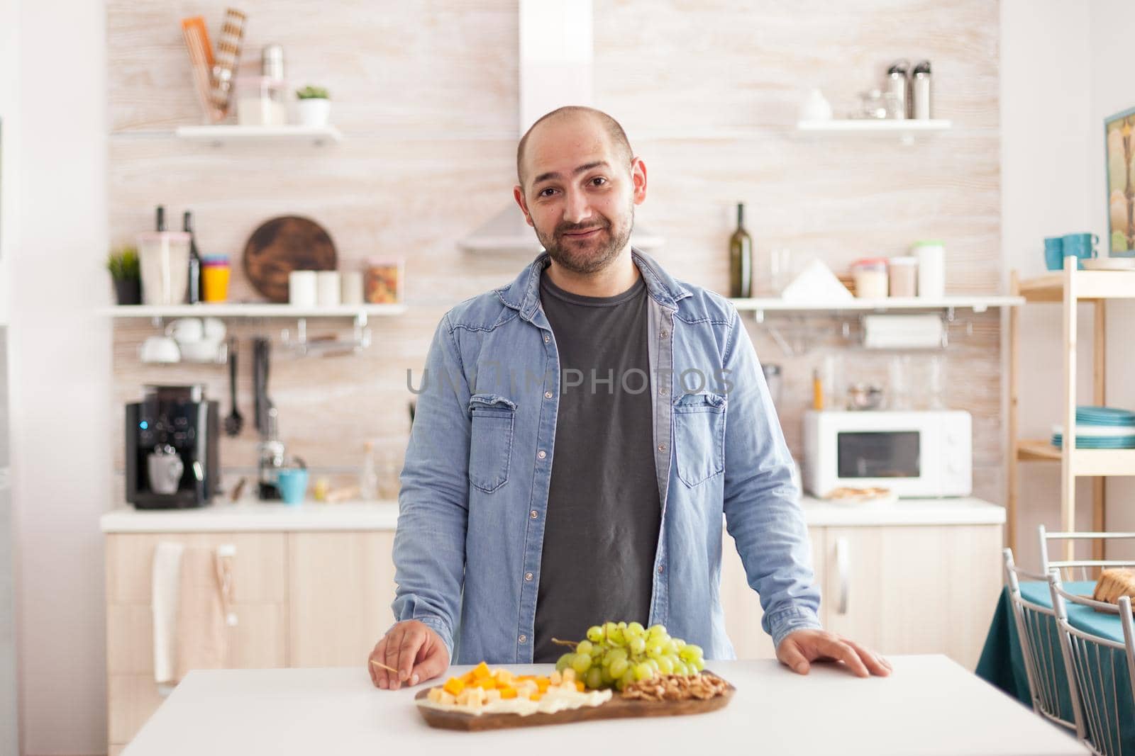 Guy in kitchen looking at camera with delicious grapes and different types of cheese on wooden board. Happy man after trying tasty cottage cheese.