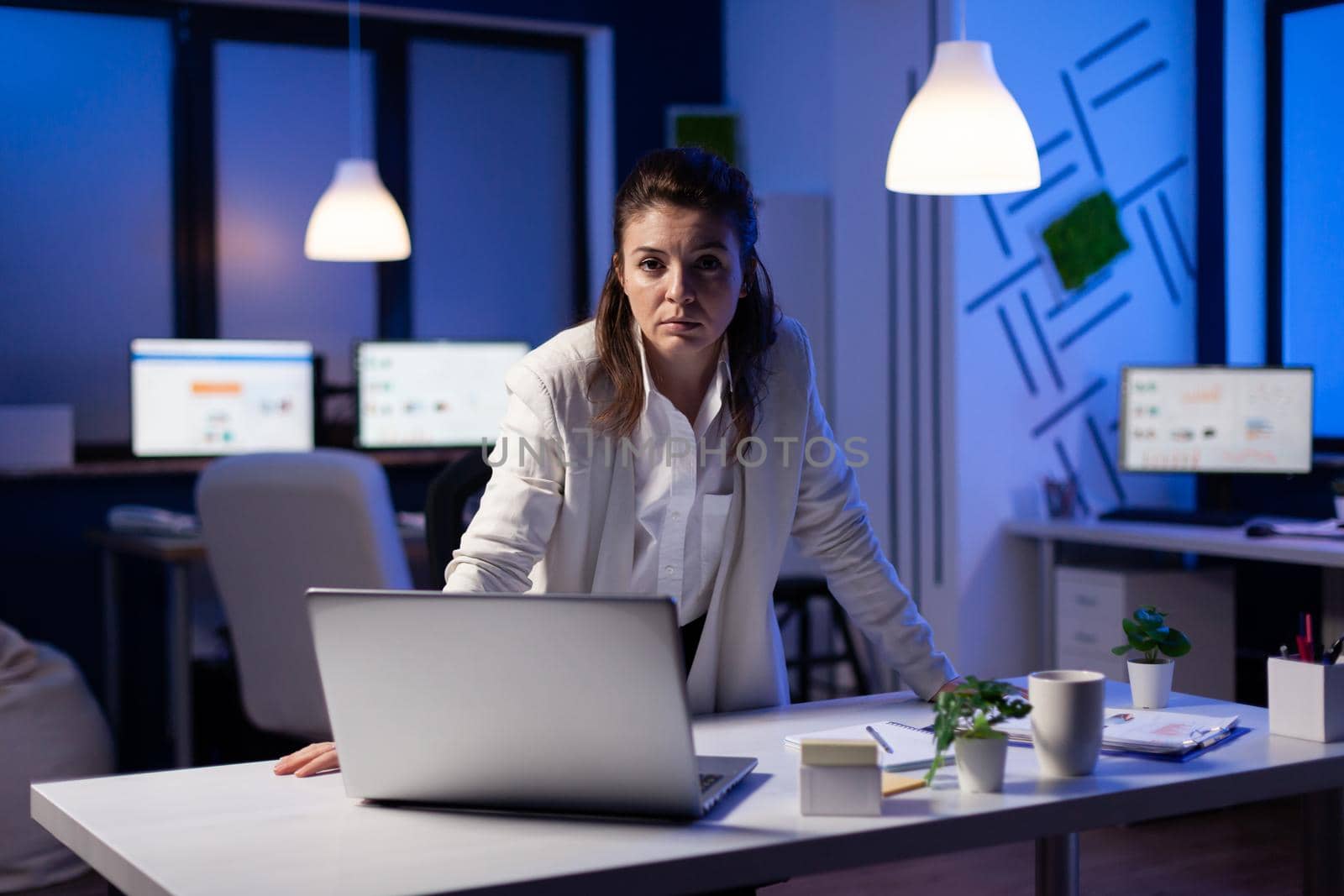 Business woman looking tired at camera standing near desk in start-up business company late at night. Focused entrepreneur using technology network wireless for developing new marketing system