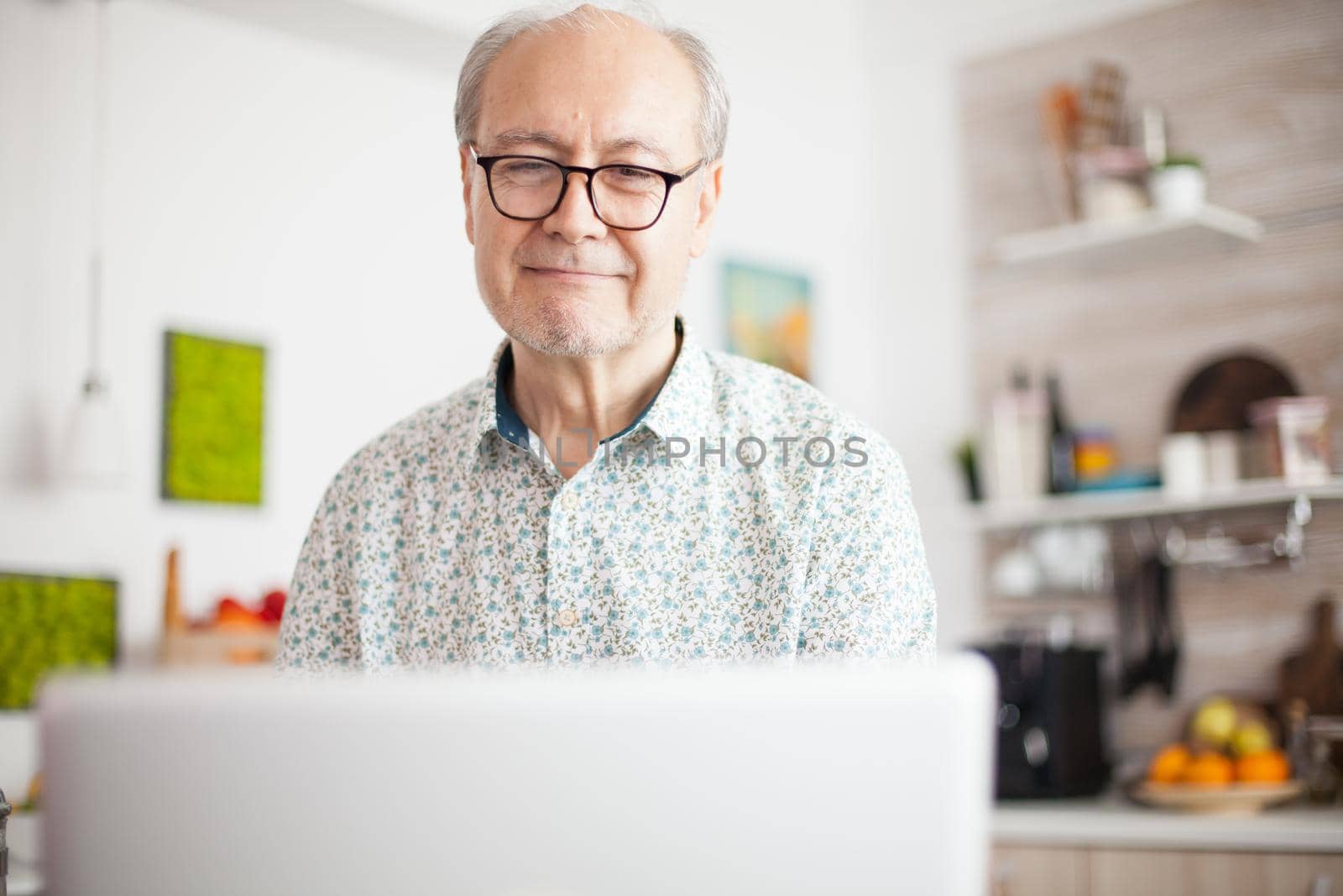 Retired man smiling while watching a movie on the laptop. Daily life of senior man in kitchen during breakfast using laptop holding a cup of coffee. Elderly retired person working from home, telecommuting using remote internet job online communication.