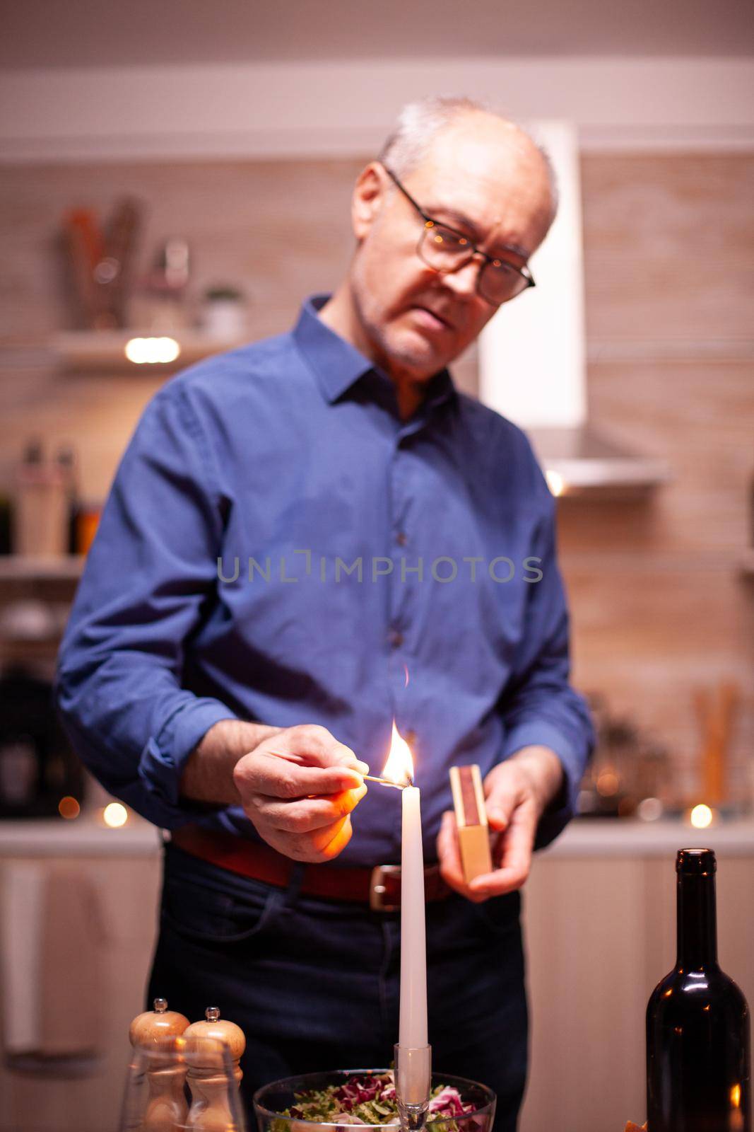 Happy senior husband using matches light candles for romantic anniversary with wife. Elderly old husband preparing festive meal with healty food for anniversary celebration, sitting near the table.