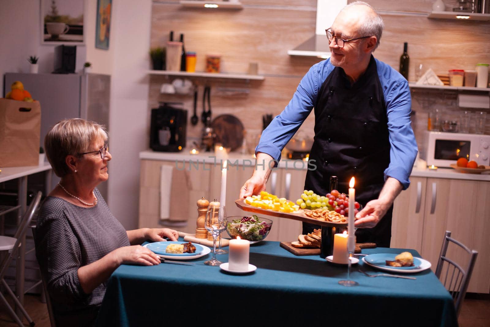 Senior man serving grapes his wife during romantic dinner in kitchen for relationship anniversary Elderly old couple talking, sitting at the dining table enjoying the meal,