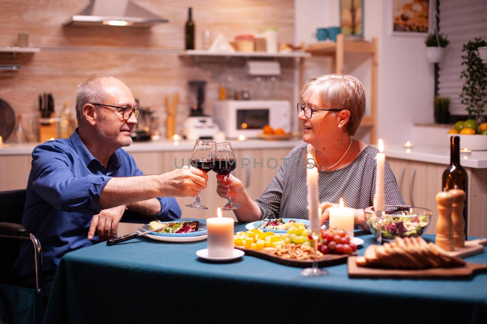 Man in wheelchair dining with wife and toasting using glasses with red wine. Wheelchair immobilized paralyzed handicapped man dining with wife at home, enjoying the meal