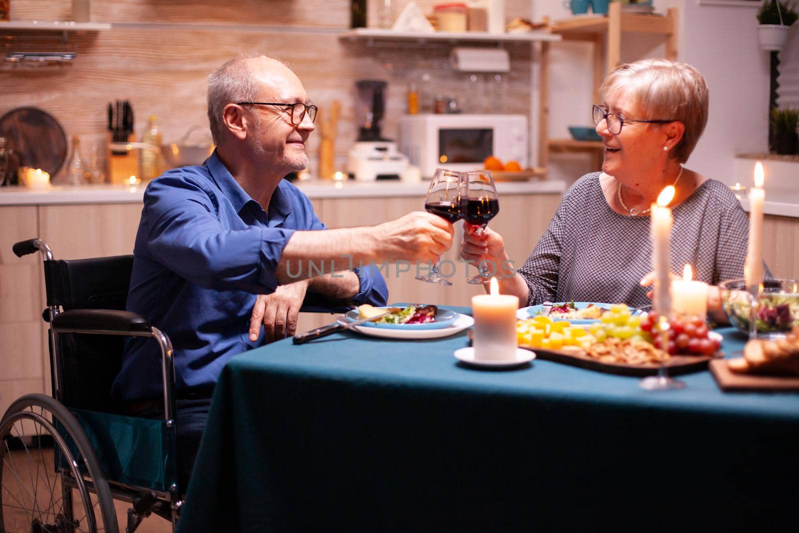 Disabled man smiling at wife while having dinner in kitchen. Wheelchair immobilized paralyzed handicapped man dining with wife at home, enjoying the meal.