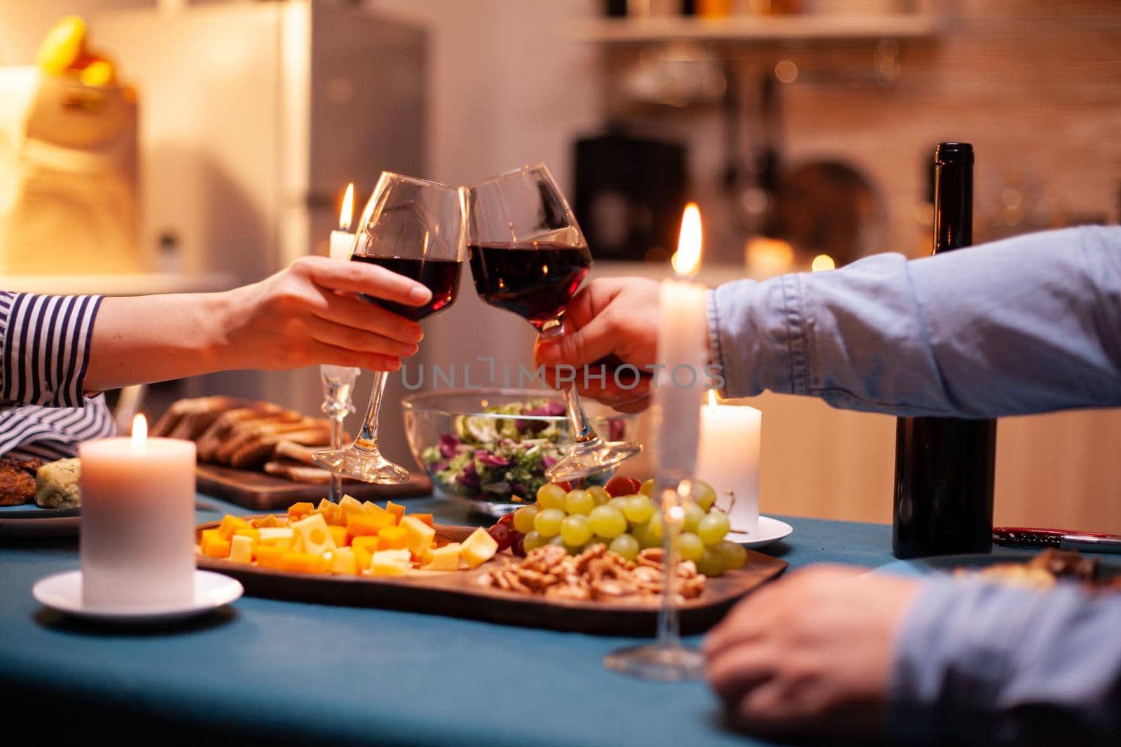Couple holding glasses with red wine during romantic relationshiop celebration. Happy cheerful young couple dining together in the cozy kitchen, enjoying the meal, celebrating anniversary romantic toast.