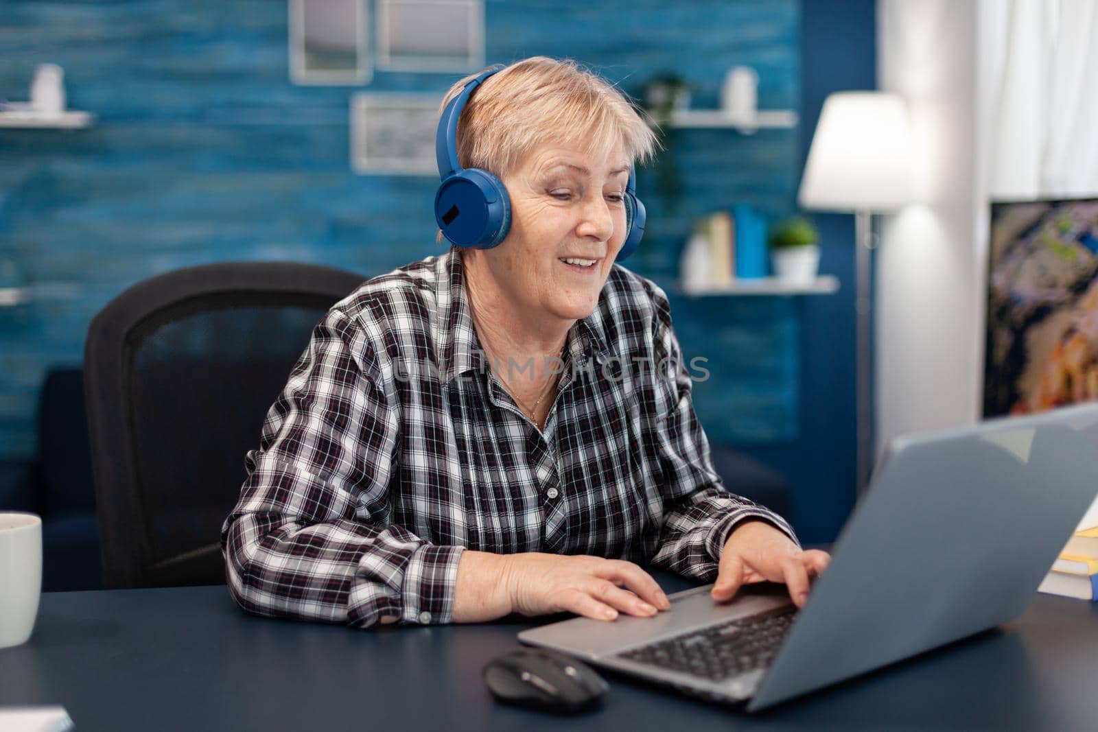 Mature lady listening music smiling and entertaining herself Retired senior woman wearing headphones in the course of using laptop computer in home living room.