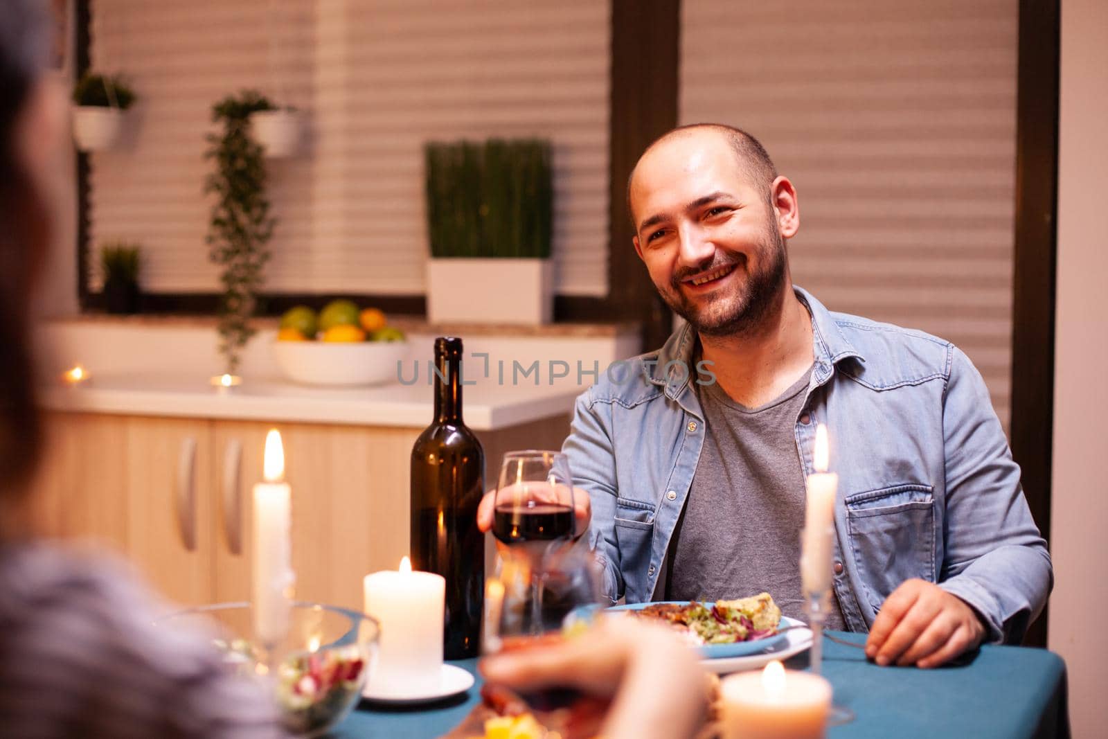 Boyfriend smiling at wife enjoying anniversary celebration in kitchen. Talking happy sitting at table dining room, enjoying the meal at home having romantic time at candle lights.