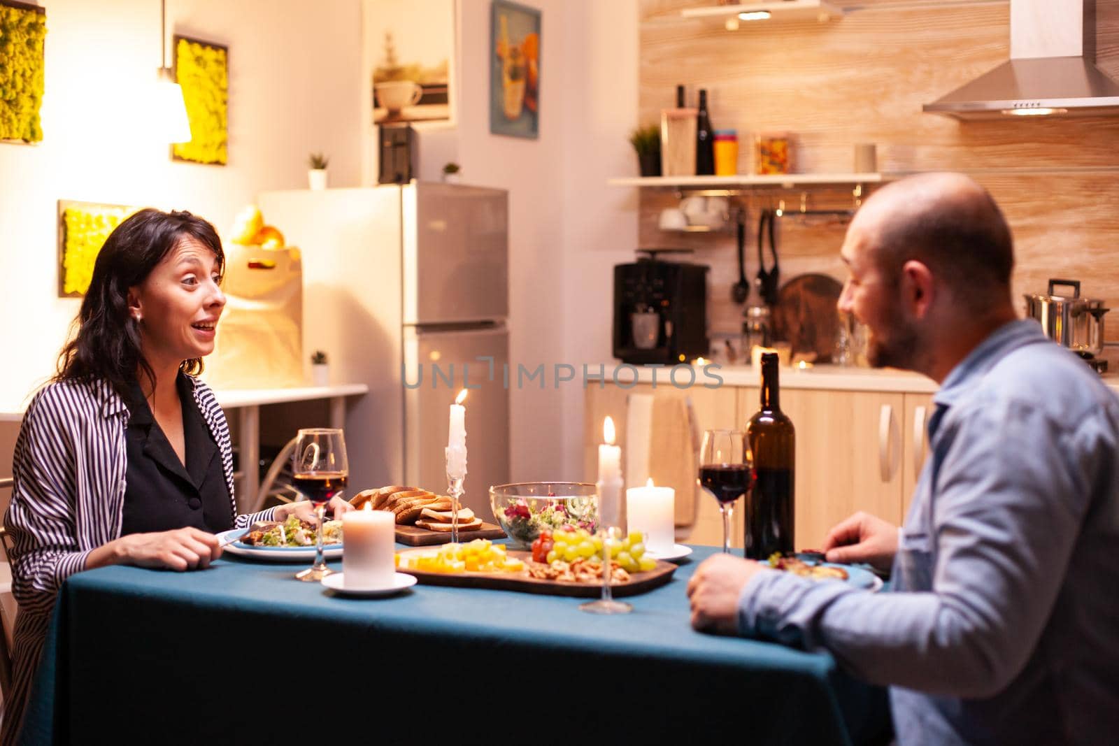 Wife looking surprised at husband during romantic dinner in kitchen. Talking happy sitting at table dining room, enjoying the meal at home having romantic time at candle lights.