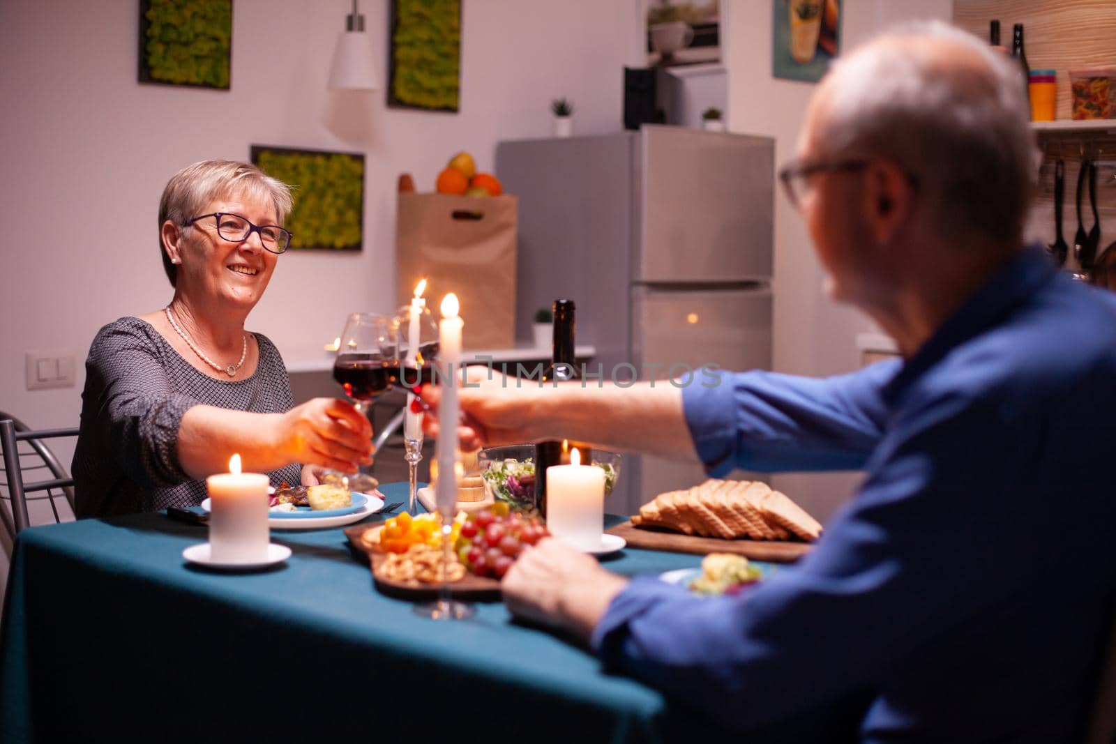 Happy pensioner couple toasting with red wine glasses in kitchen. Senior couple sitting at the table in kitchen, talking, enjoying the meal, celebrating their anniversary in the dining room.