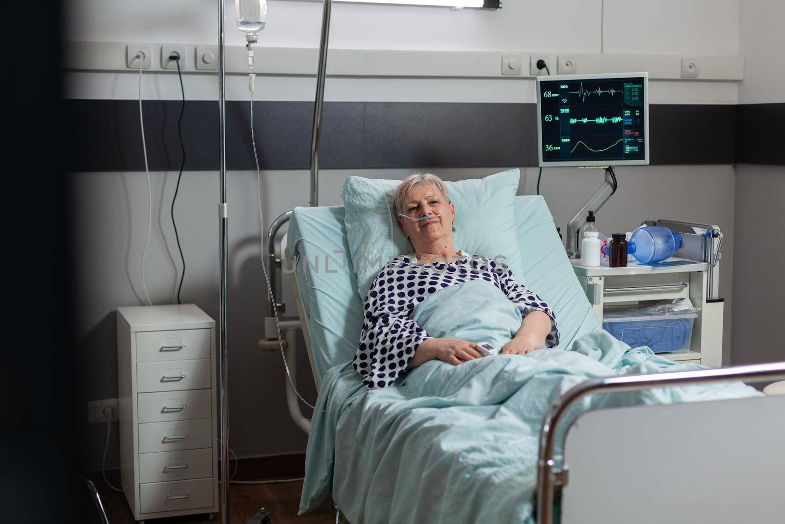 Portrait of senior woman smiling looking at camera laying in hospital bed betting treatment through iv drip bag. Breathing with help from oxygen mask during illness recovery.