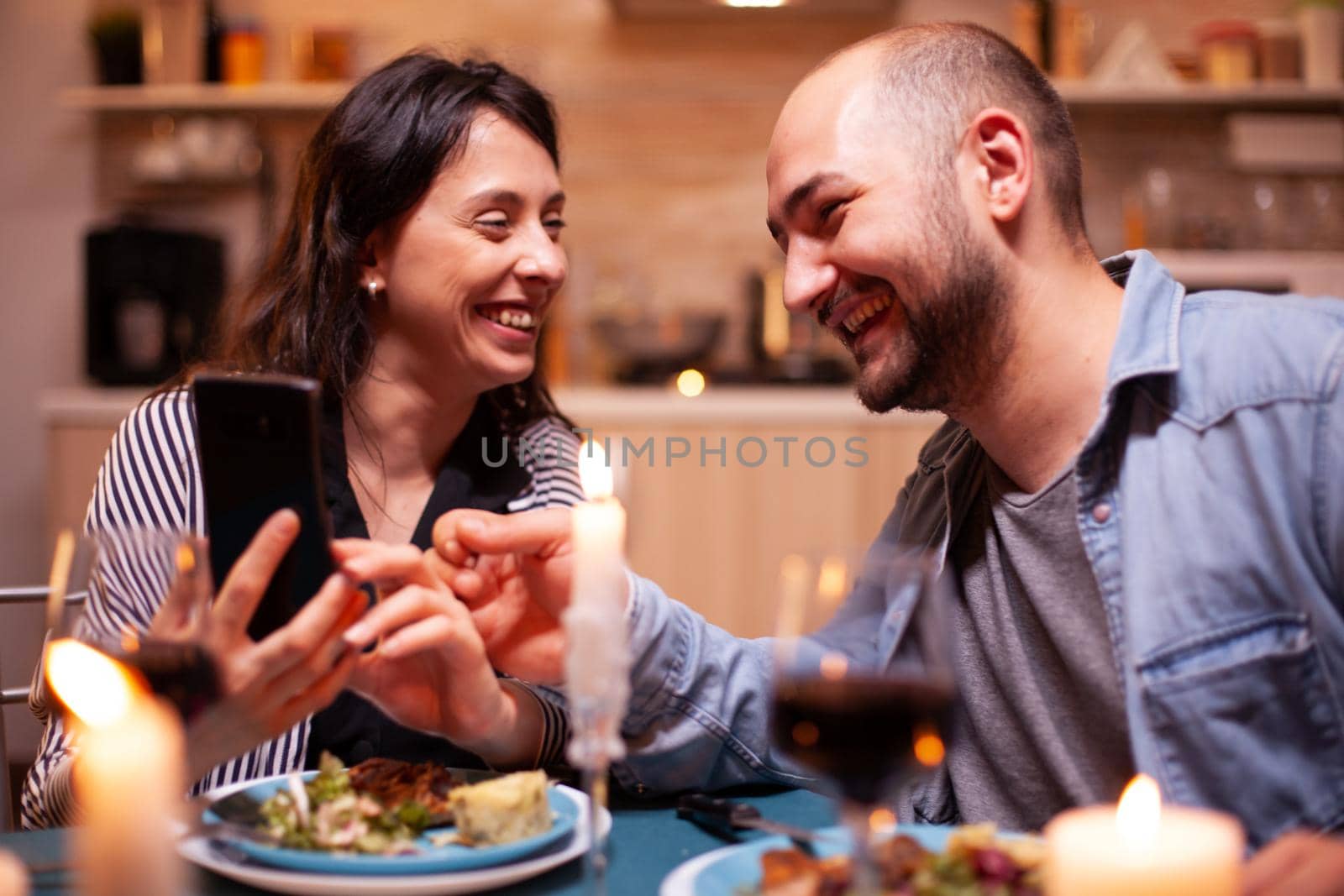 Cheerful couple laughing using smartphone during relationship anniversary. Adults sitting at the table in the kitchen browsing, searching, using smartphones, internet, celebrating .