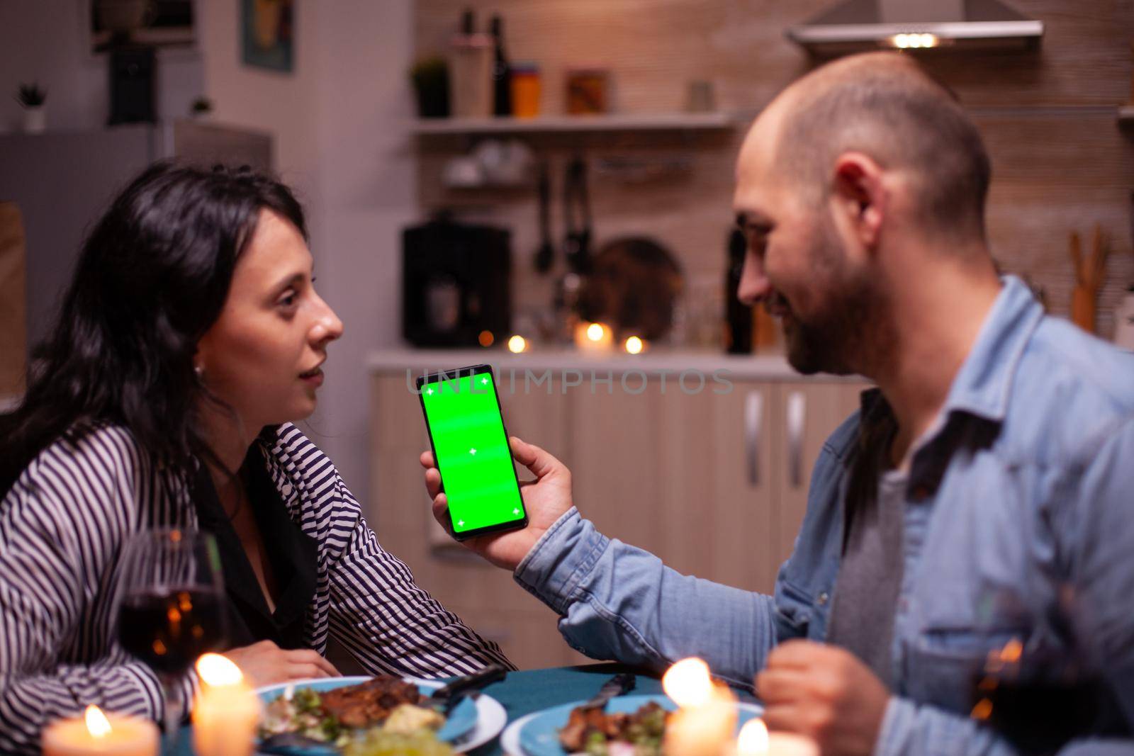 Guy holding phone with green screen while having romantic dinner with wife. Happy looking at green screen template chroma key isolated smartphone display using technology internet.