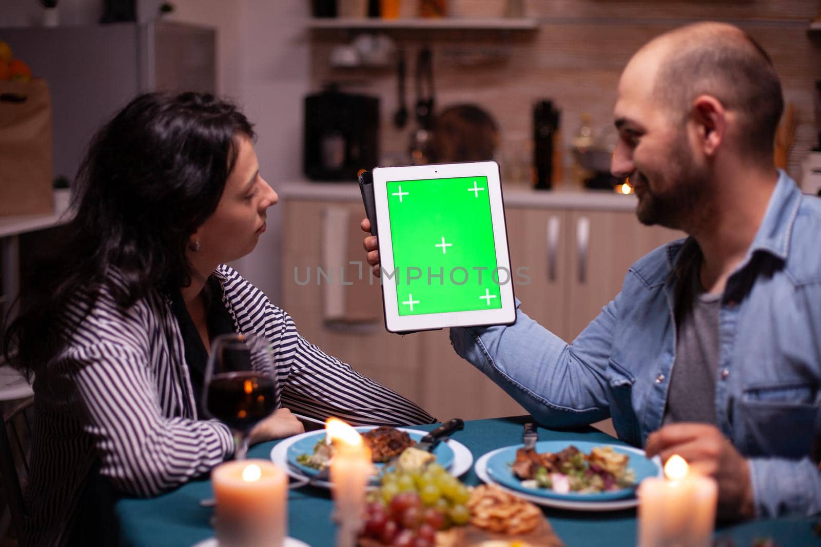 Husband holding tablet pc with green mock up while enjoying romantic dinner with wife. Man and woman looking at green screen template chroma key display sitting at the table in kitchen during dinner.
