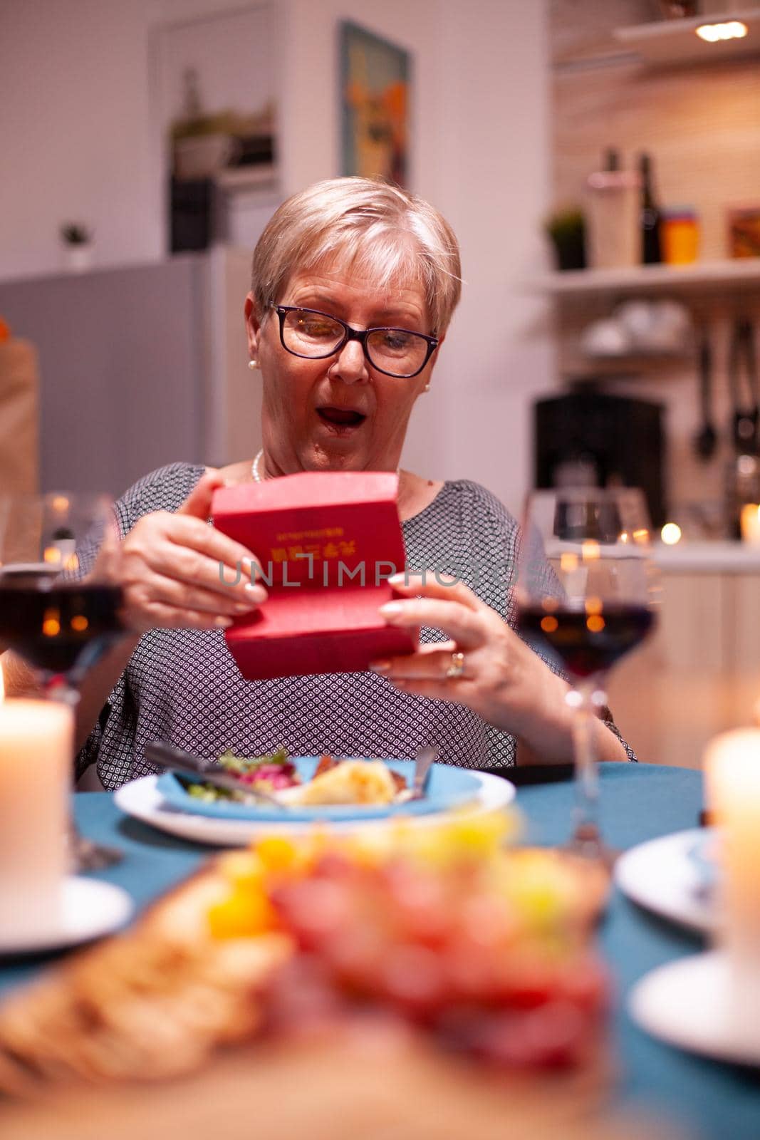Elderly woman looking shocked at gift box during festive dinner. Happy cheerful elderly couple dining together at home, enjoying the meal, celebrating their marriage , surprise holiday
