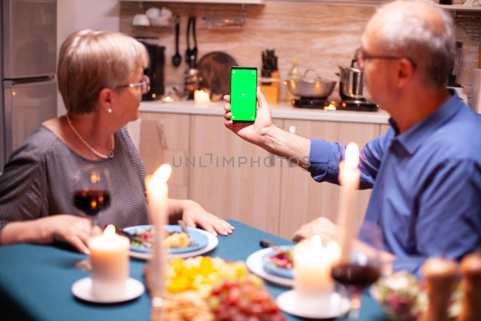 Old retired senior couple holding mockup phone at dinner. Aged people looking at mockup template chroma key isolated smart phone display using techology internet sitting at the table in kitchen.