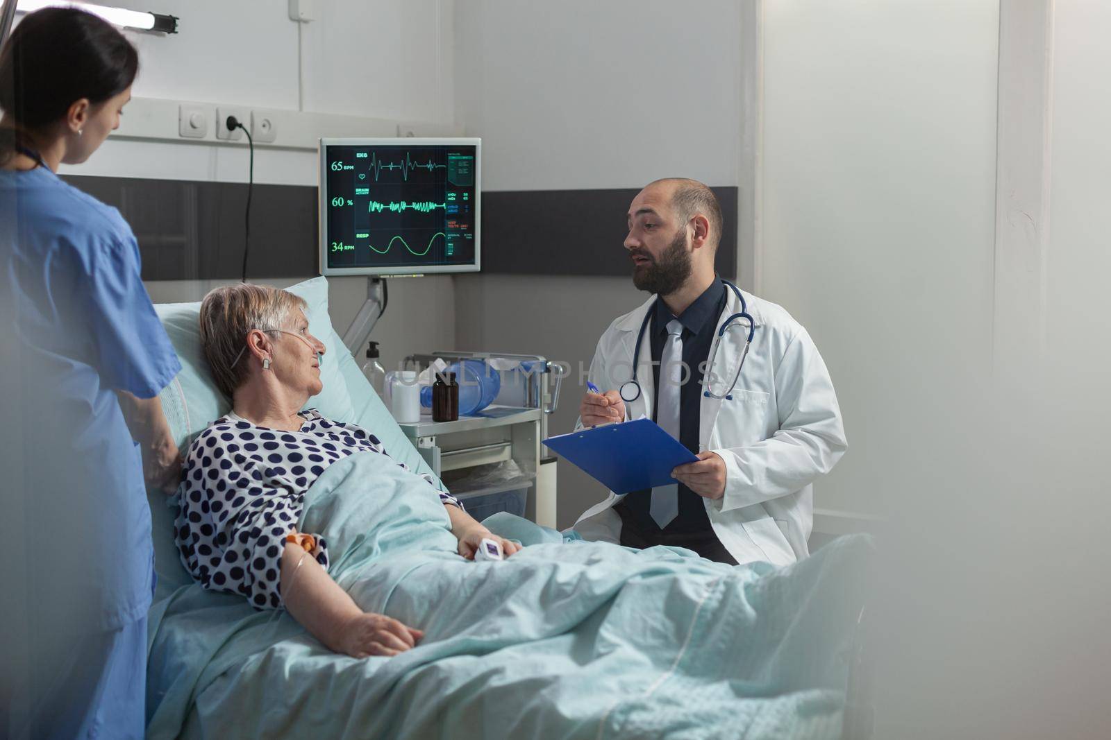 Doctor talking with elderly patient sitting next to bed in hospital room, giving expertise for treatment. Senior woman breathing with help from oxygen mask laying in bed.