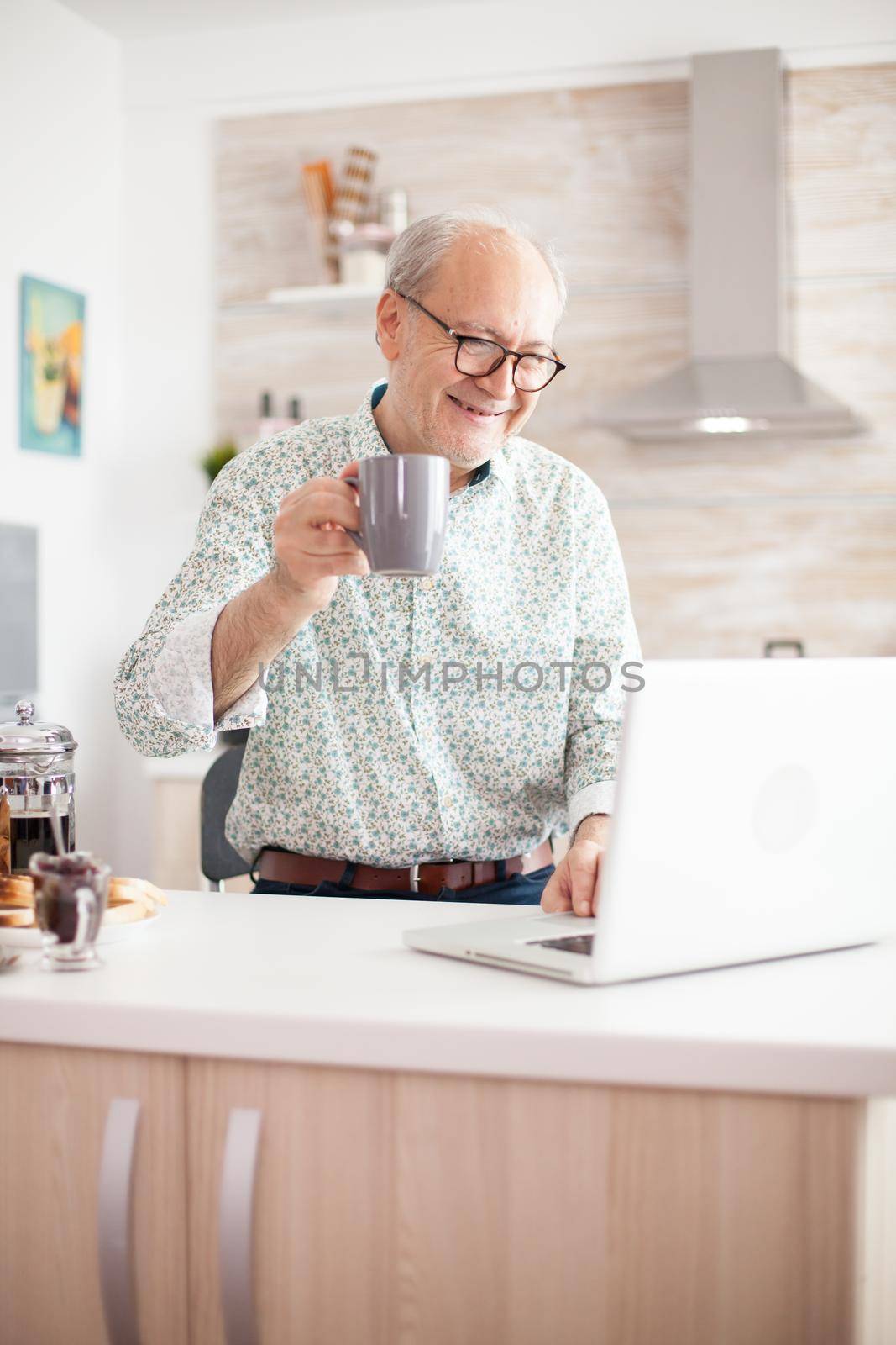 Cheerful senior man during video conference in kitchen on the laptop while enjoying breakfast and a cup of coffee. Elderly person using internet online chat technology video webcam making a video call connection camera communication conference call