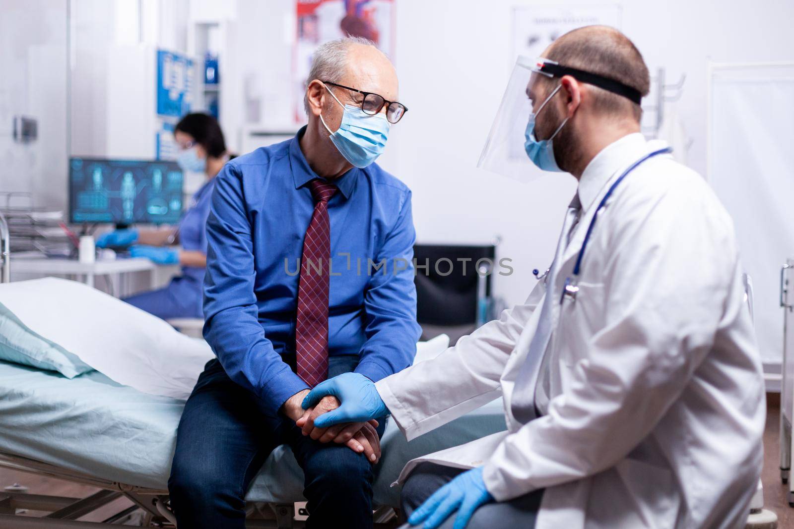 Doctor giving compassion to senior man wearing protection mask against coronavirus during consultation in hospital examination room.