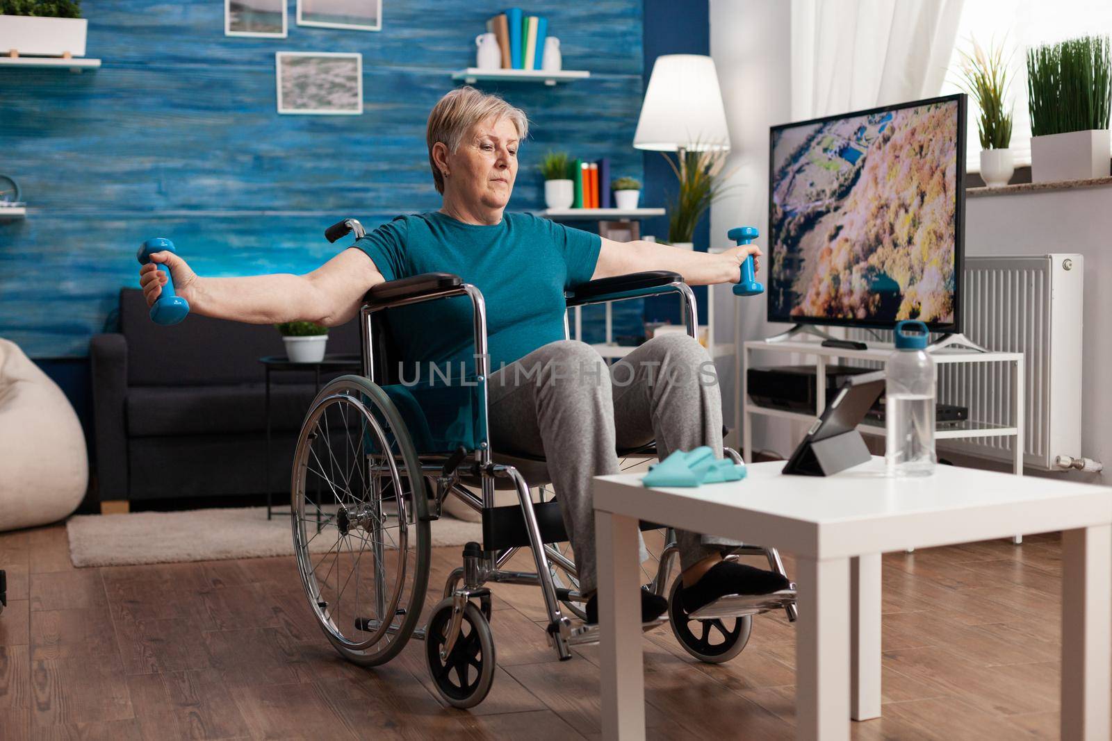 Handicapped senior woman in wheelchair stretching arms muscles exercising body resistance using workout dumbbells in living room. Handicapped pensioner looking at lifestyle aerobics video on tablet