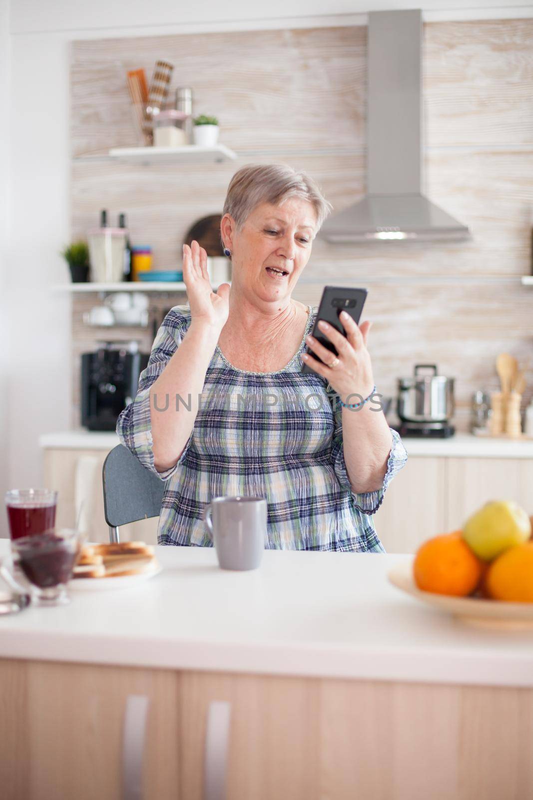 Retired woman saying hello while having a video call with family using smartphone in kitchen during breakfast. Elderly person using internet online chat technology video webcam making video call connection camera communication conference call
