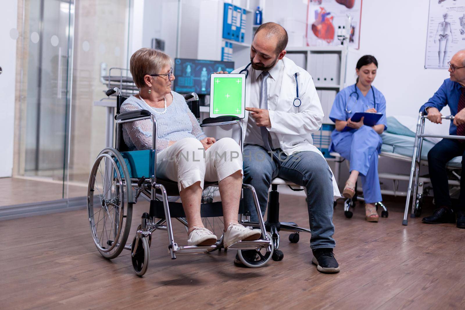 Doctor pointing at green mockup on tablet pc while telling senior woman in wheelchair about her rehabilitation. Man with disabilities ,walking frame sitting in hospital bed. Health care system,