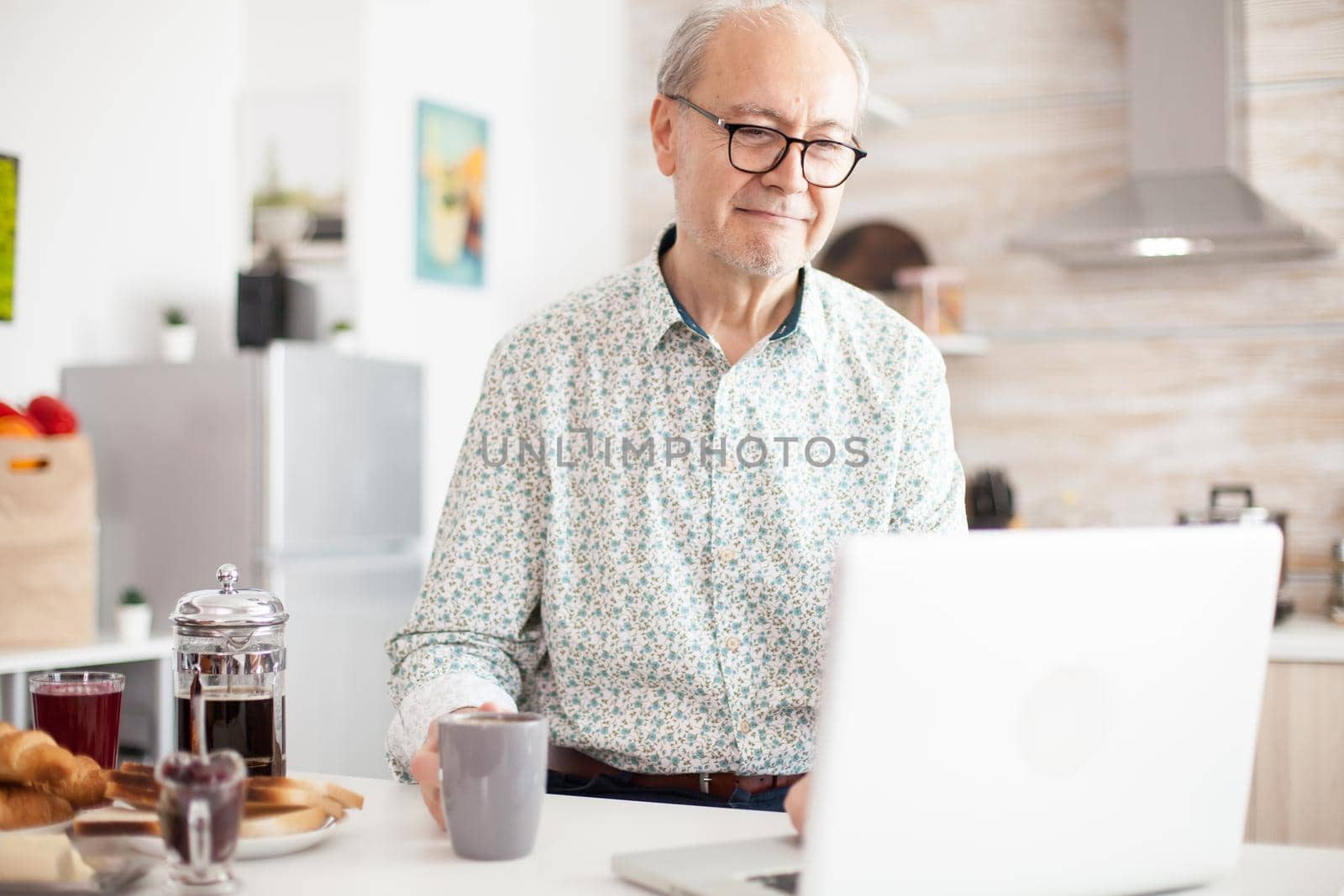 Old man in the kitchen working remotely. Daily life of senior man in kitchen during breakfast using laptop holding a cup of coffee. Elderly retired person working from home, telecommuting using remote internet job online communication on modern technology notebook