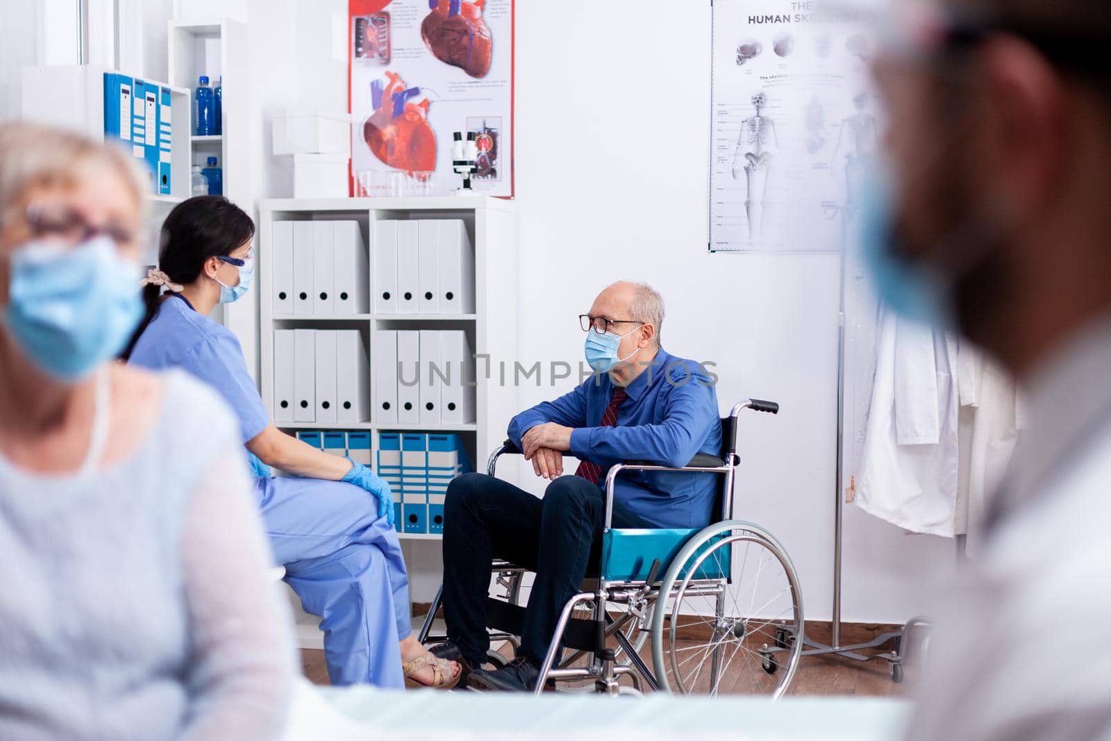 Disabled old man sitting in wheelchair wearing face mask agasint coronavirus while talking with medical staff in hospital room. Global health crisis, medical system during pandemic, sick elderly.