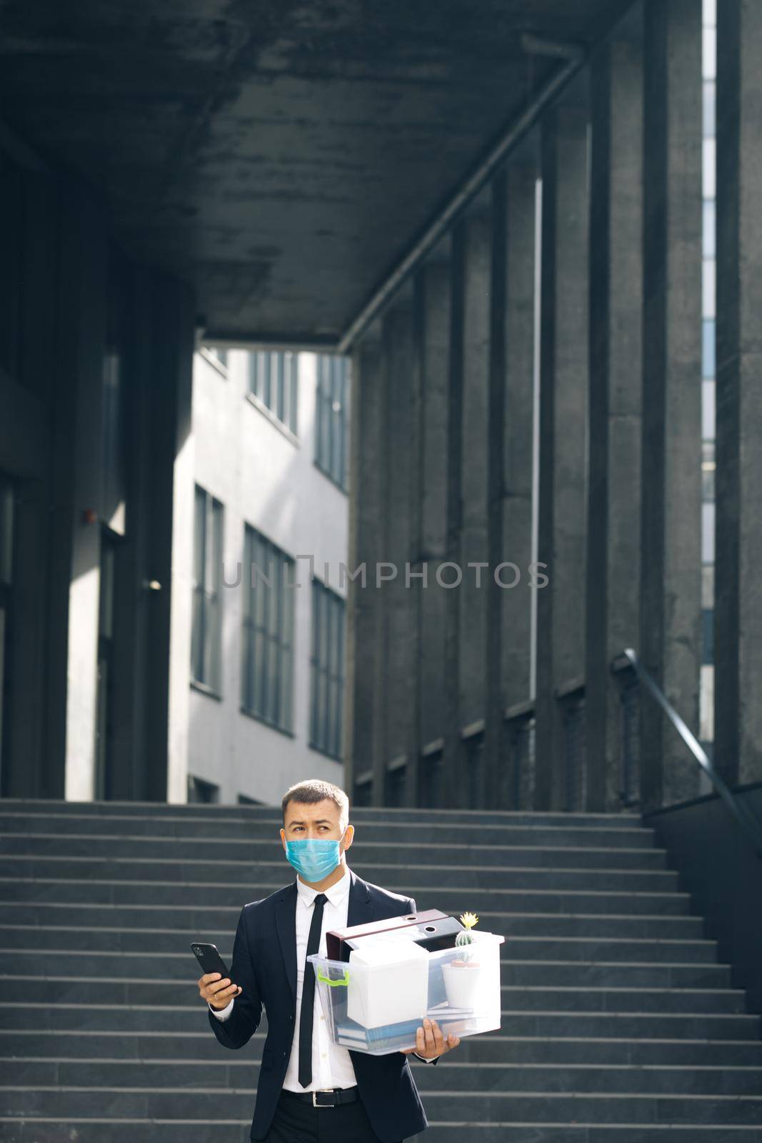 Businessman in medical mask with box of personal stuff walking the street use phone. Finance and industry. Fired man lost job. Business style suit. Coronavirus outdoors social distancing.