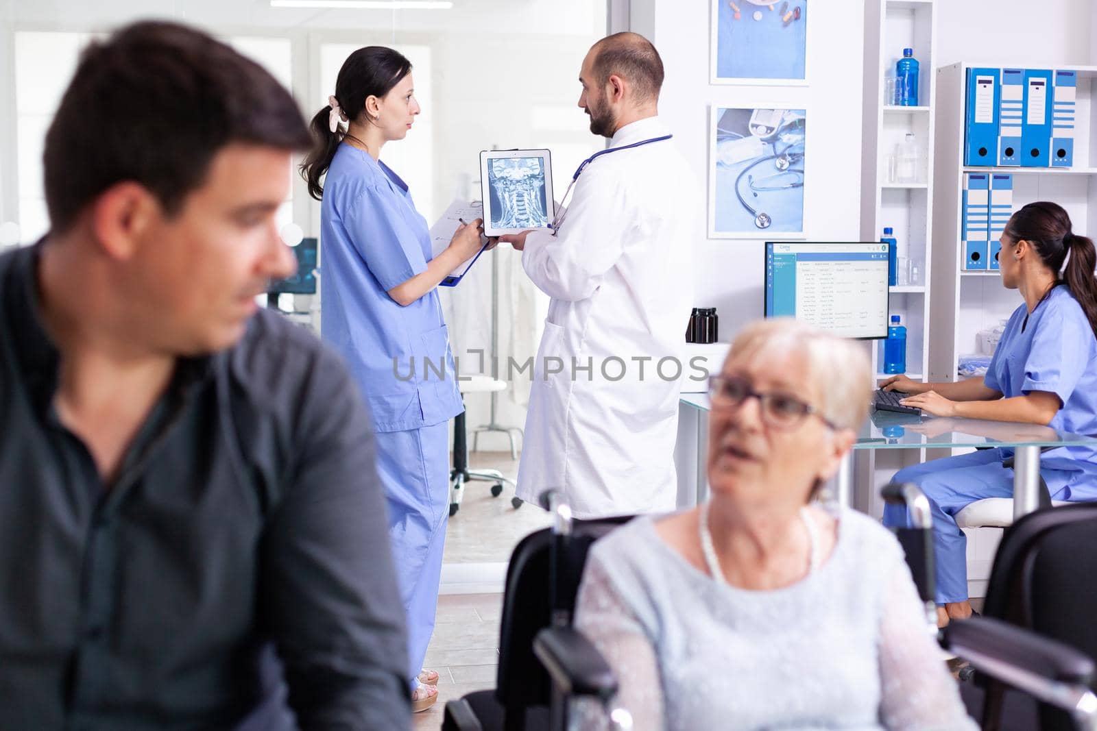 Medical staff holding tablet pc with patient radiography in hospital hallway. Doctor discussing diagnosis with nurse in clinic waiting area. Old woman sitting in wheelchair.