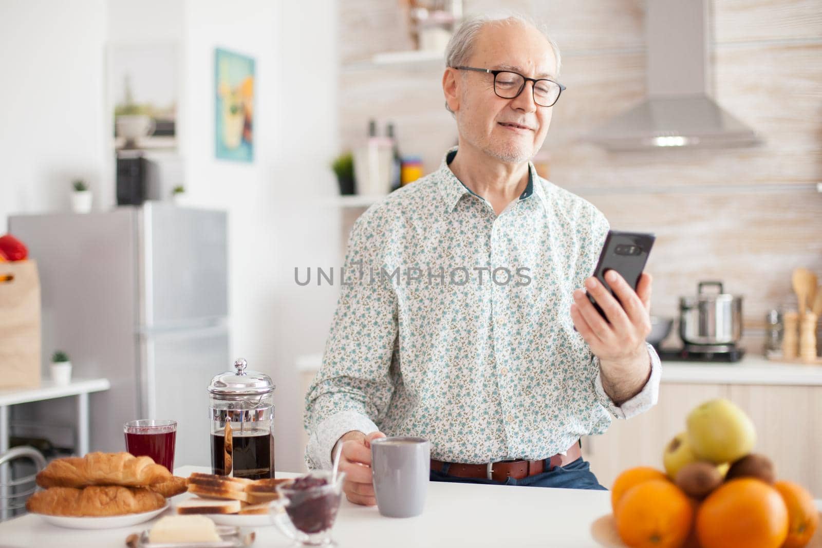 Senior man browsing on internet using smartphone in kitchen while enjoying morning coffee during breakfast. Authentic portrait of retired senior enjoying modern internet online technology