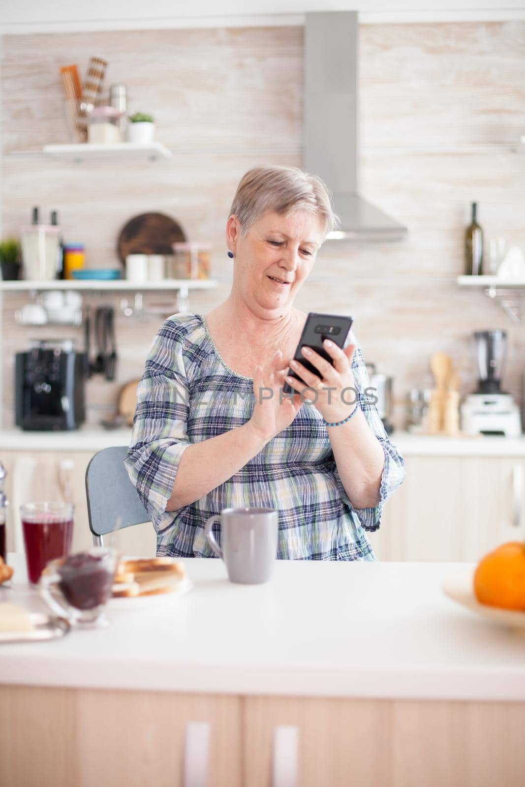 Elderly woman reading message on smartphone during breakfast in kitchen. Authentic elderly person using modern smartphone internet technology. Online communication connected to the world, senior leisure time with gadget