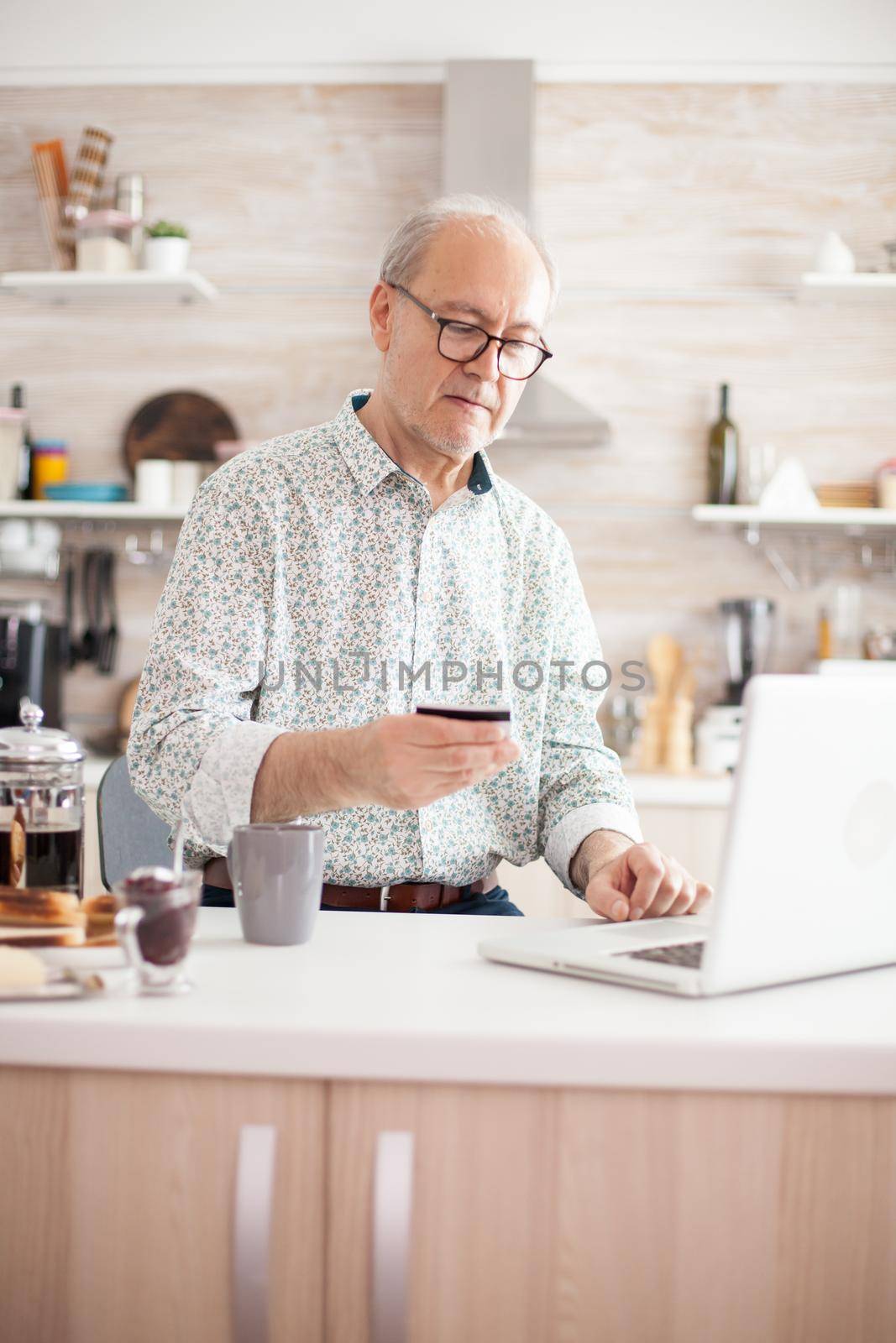 Elderly man holding credit card, using modern payment system. Pensioner paying online using credit card and application from laptop during breakfast in kitchen. Retired elderly person using internet payment home bank buying with modern technolog