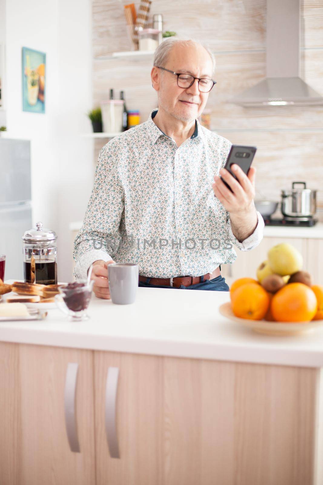 Retired old man watching video watching video on smartphone in kitchen while having breakfast. Using modern technology while enjoying morning coffee during breakfast. Authentic portrait of retired senior internet online technology