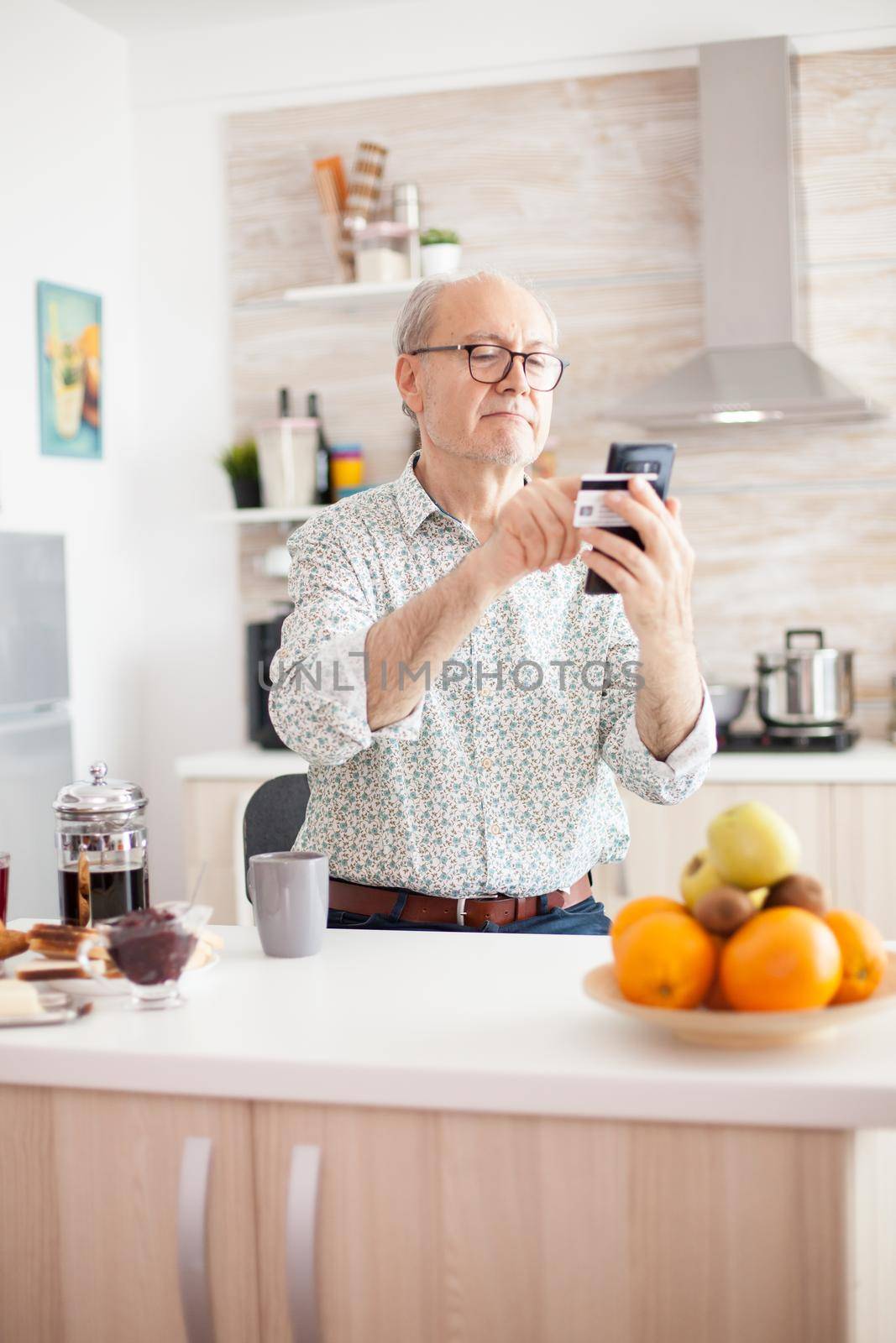 Senior man shopping online using smartphone and holding credit card during breakfast in kitchen. Retired elderly person using internet payment home bank buying with modern technology