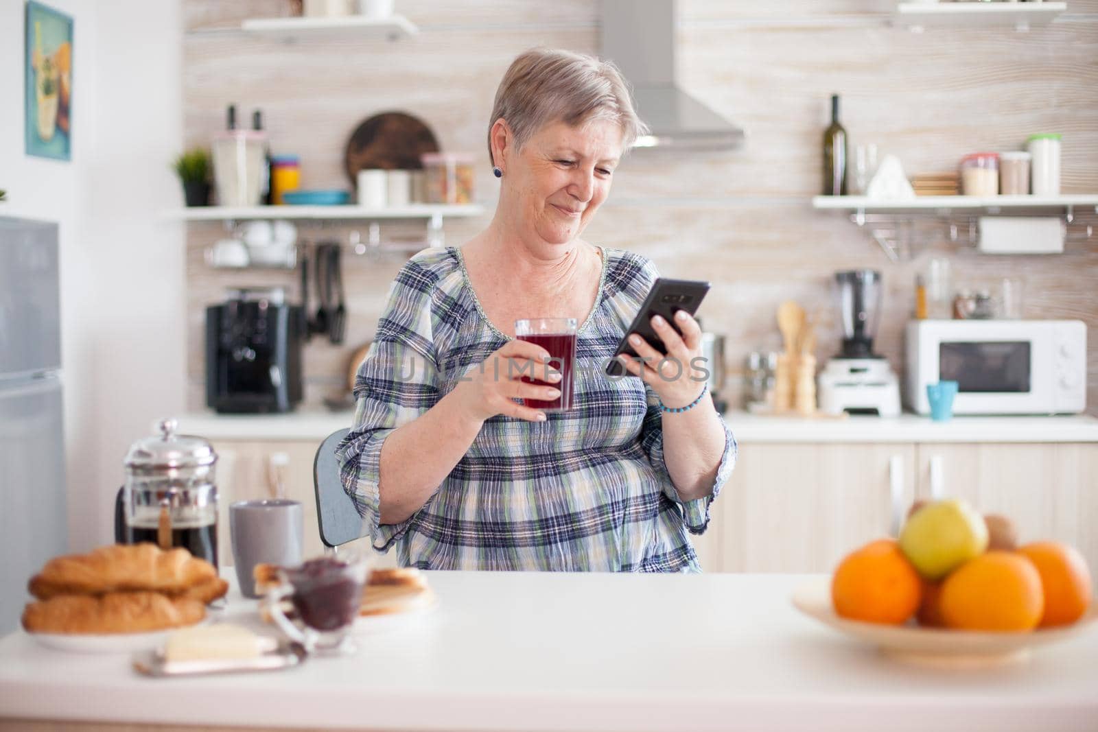 Leisure morning for senior woman using modern technology in bright cozy kitchen. Authentic elderly person using modern smartphone internet technology. Online communication connected to the world, senior leisure time with gadget at retirement age