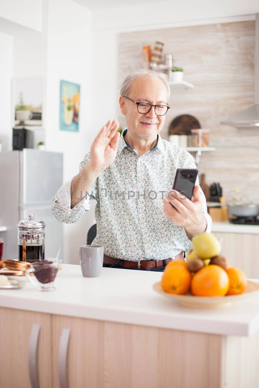 Senior man having a conversation, using his modern phone. Elderly aged man waving during a video conference with family in kitchen. Elderly person using internet online chat technology video webcam making a video call connection camera communication conference call