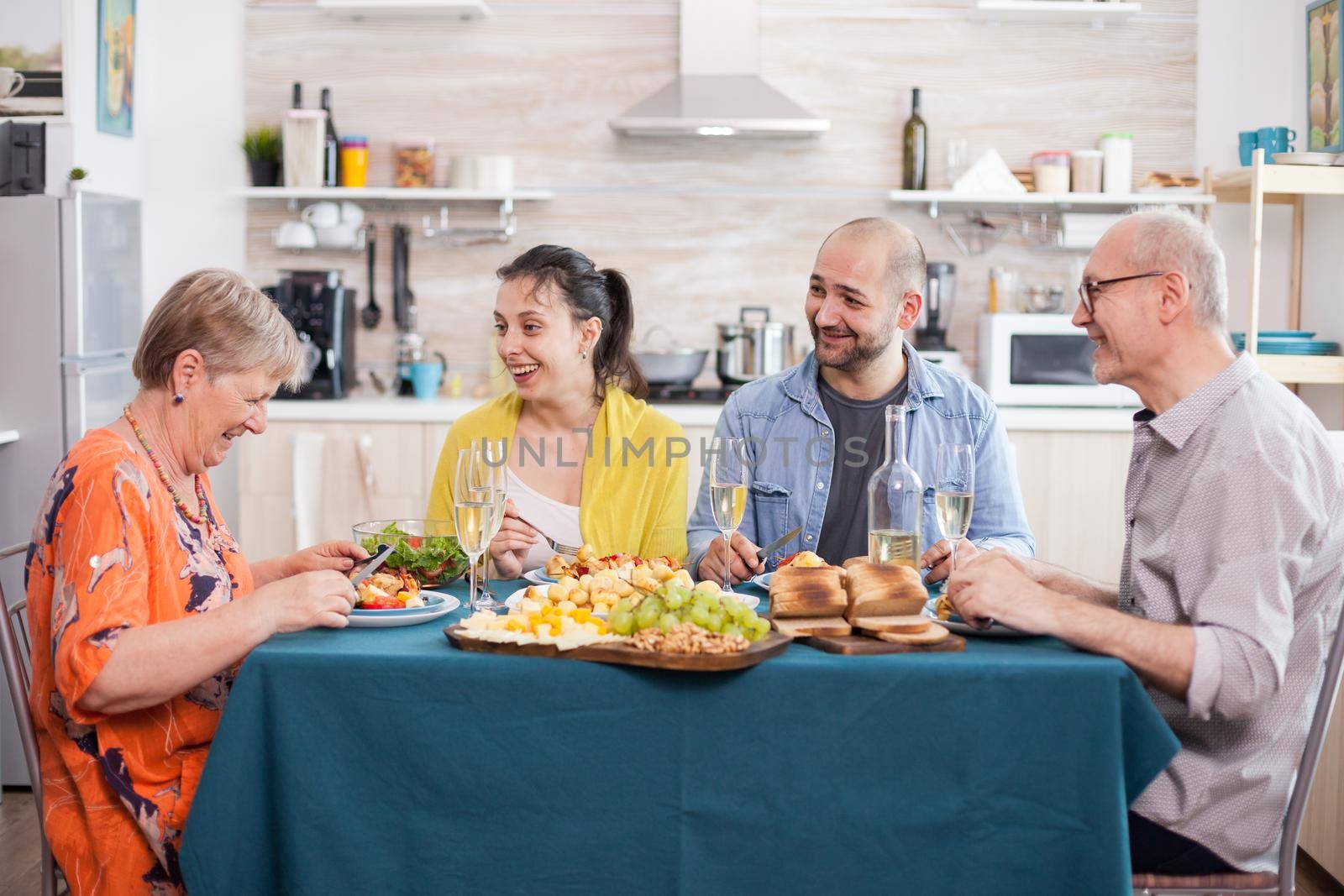 Senior woman laughing while having lunch with family in kitchen. Cheerful senior father holding wine glass. Tasty potatoes.