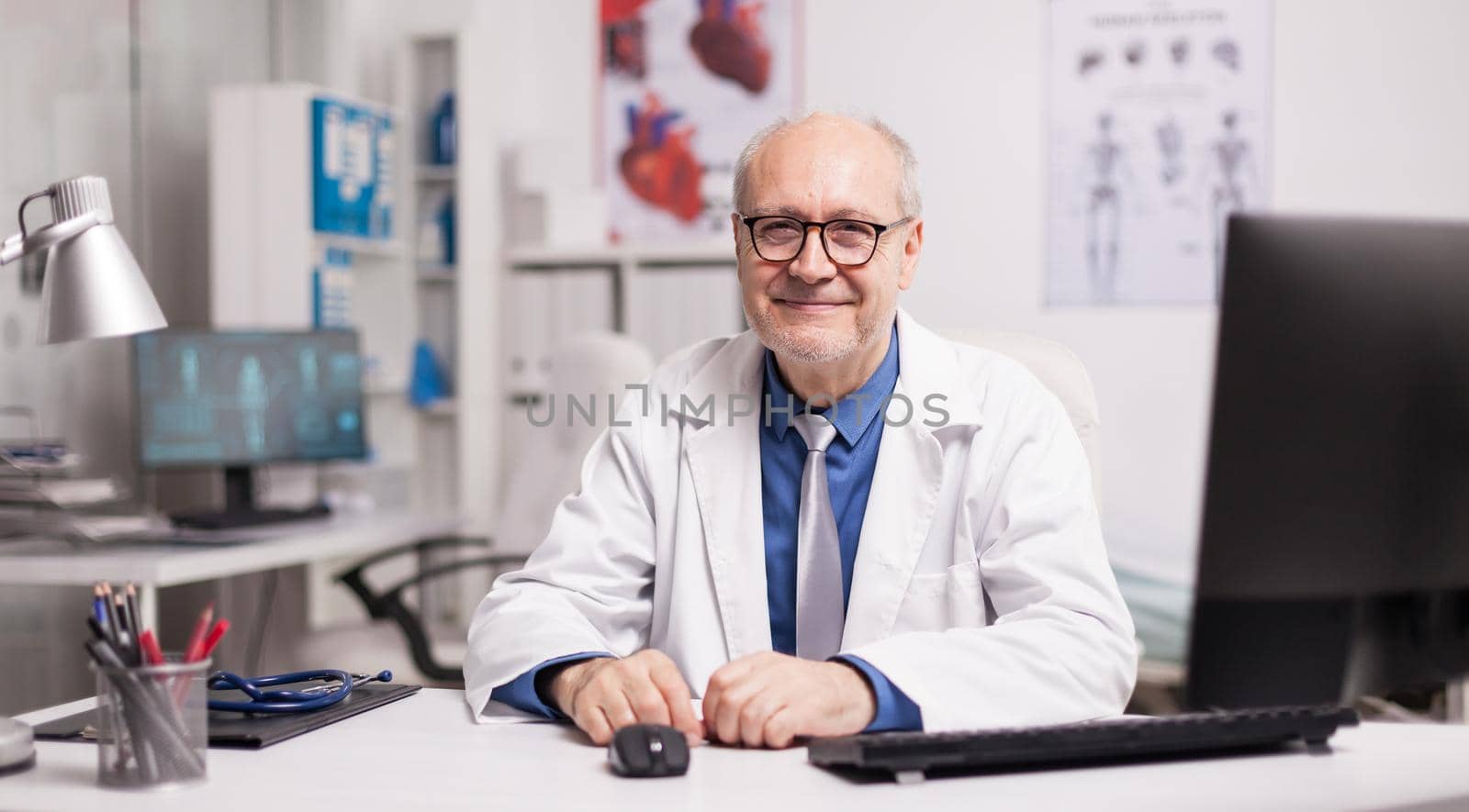 Mature doctor wearing eyeglasses and smiling in hospital cabinet sitting at desk.