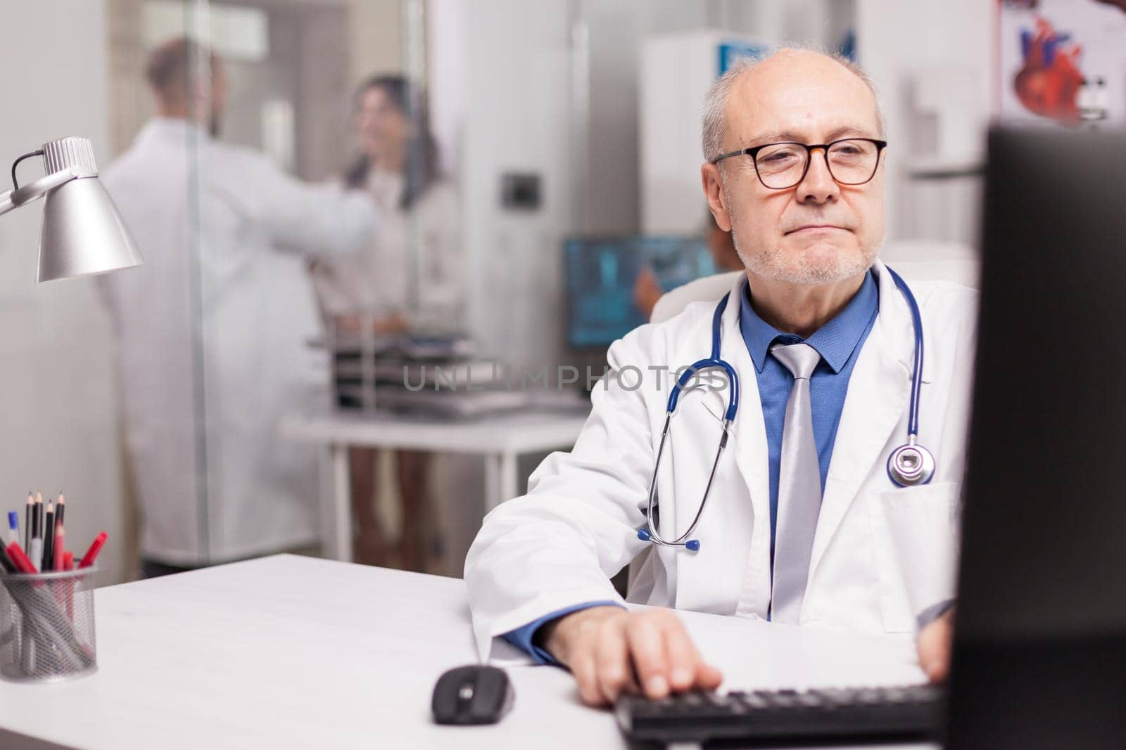 Close up of physician with grey hair typing document about new drug in hospital cabinet and young medic discussing with patient in the background wearing white coat.