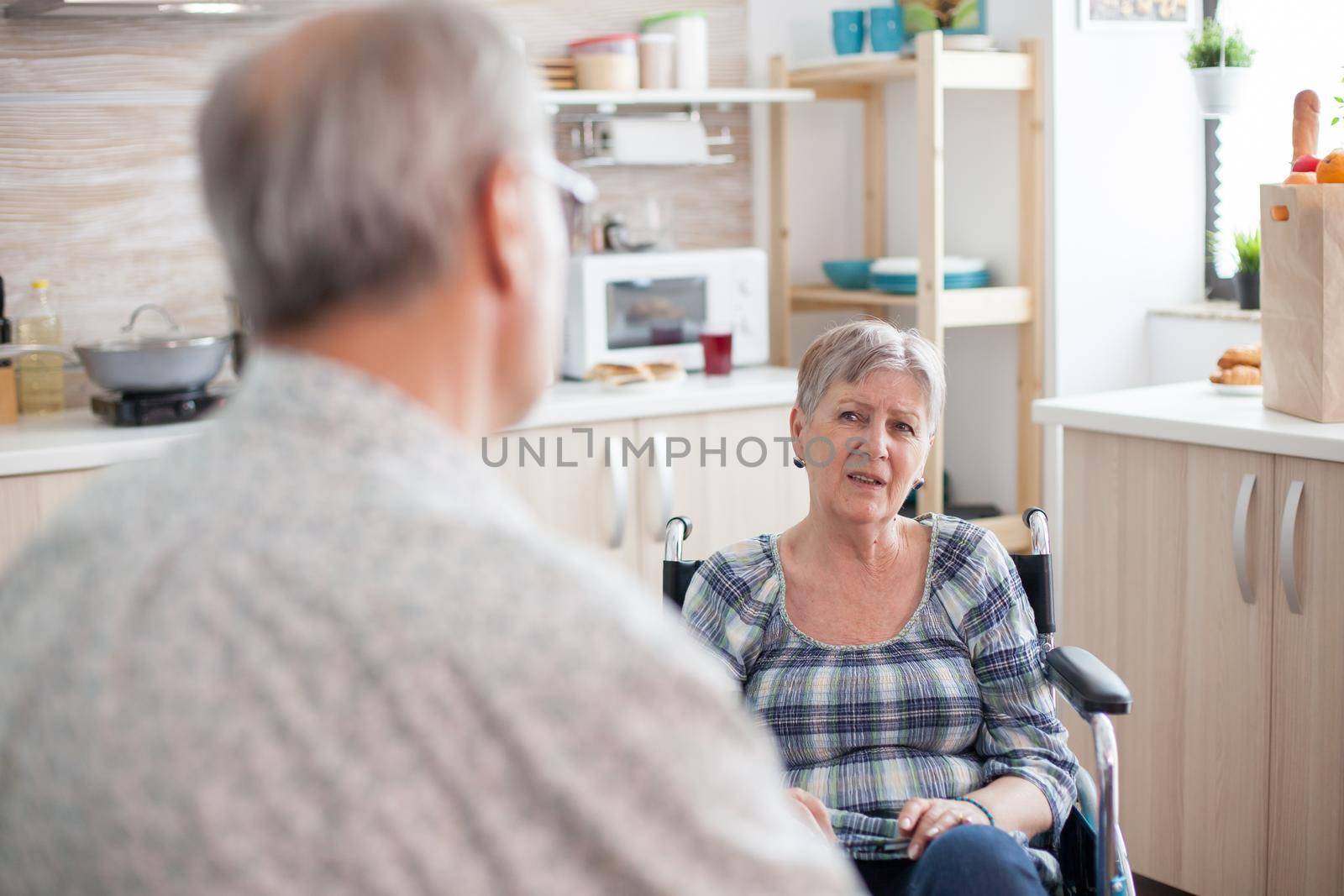 Gray Haired man chatting with paralysed wife. Retired invalid woman in wheelchair having a conversation with old elderly husband in kitchen. Old man talking with wife. Living with disabled person with walking disabilities