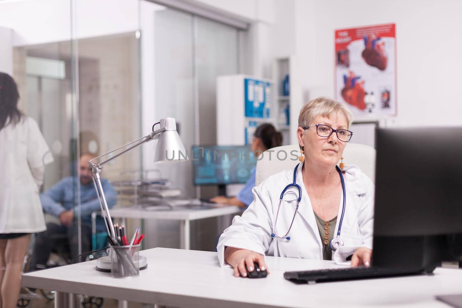 Elderly aged woman doctor working on pc in hospital office, Young therapist discussing with invalid patient in wheelchair and nurse looking at x-ray on computer screen.