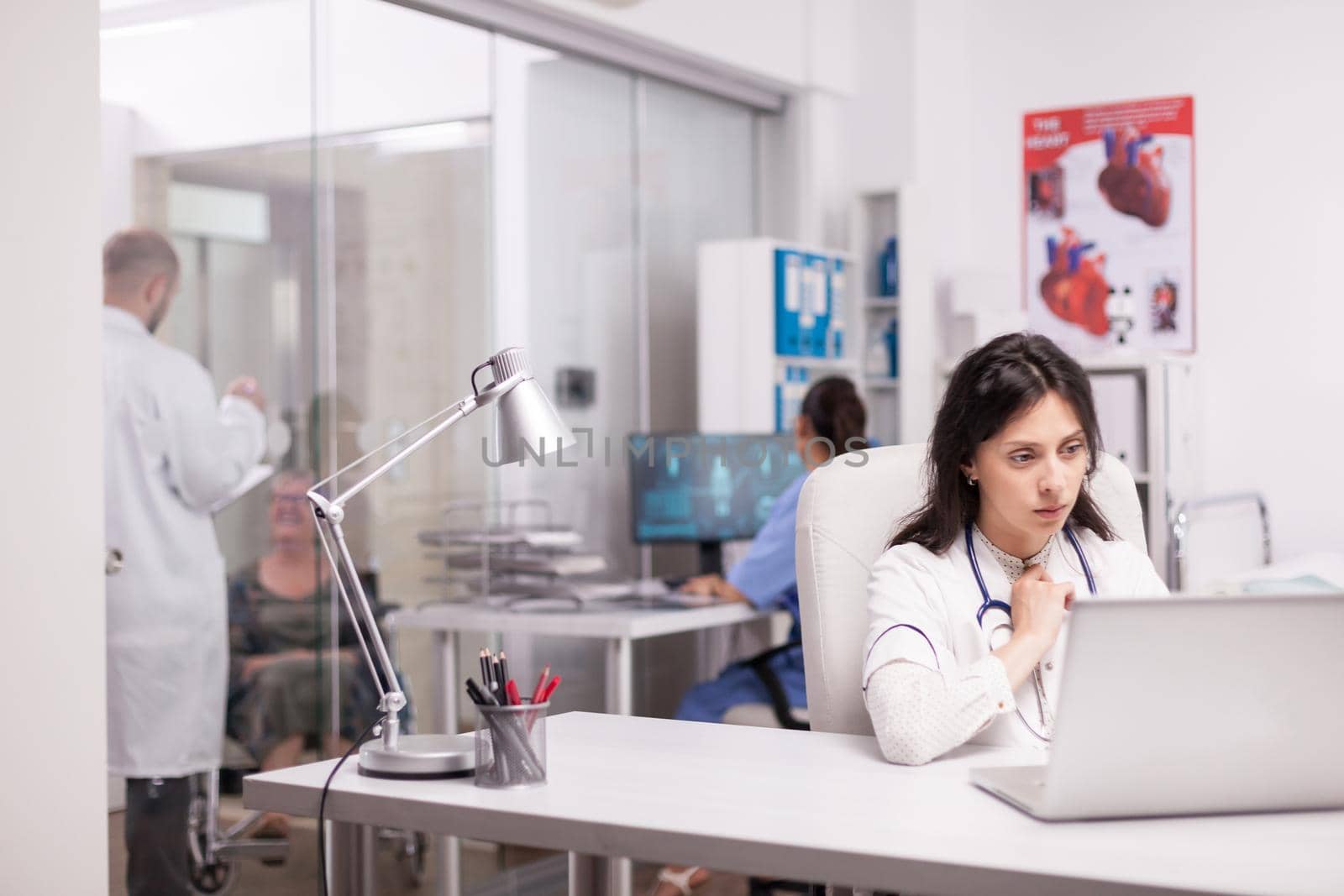 Worried young doctor working on laptop in hospital office while her colleague is talking with disabled senior woman in wheelchair on clinic corridor and nurse in blue uniform.