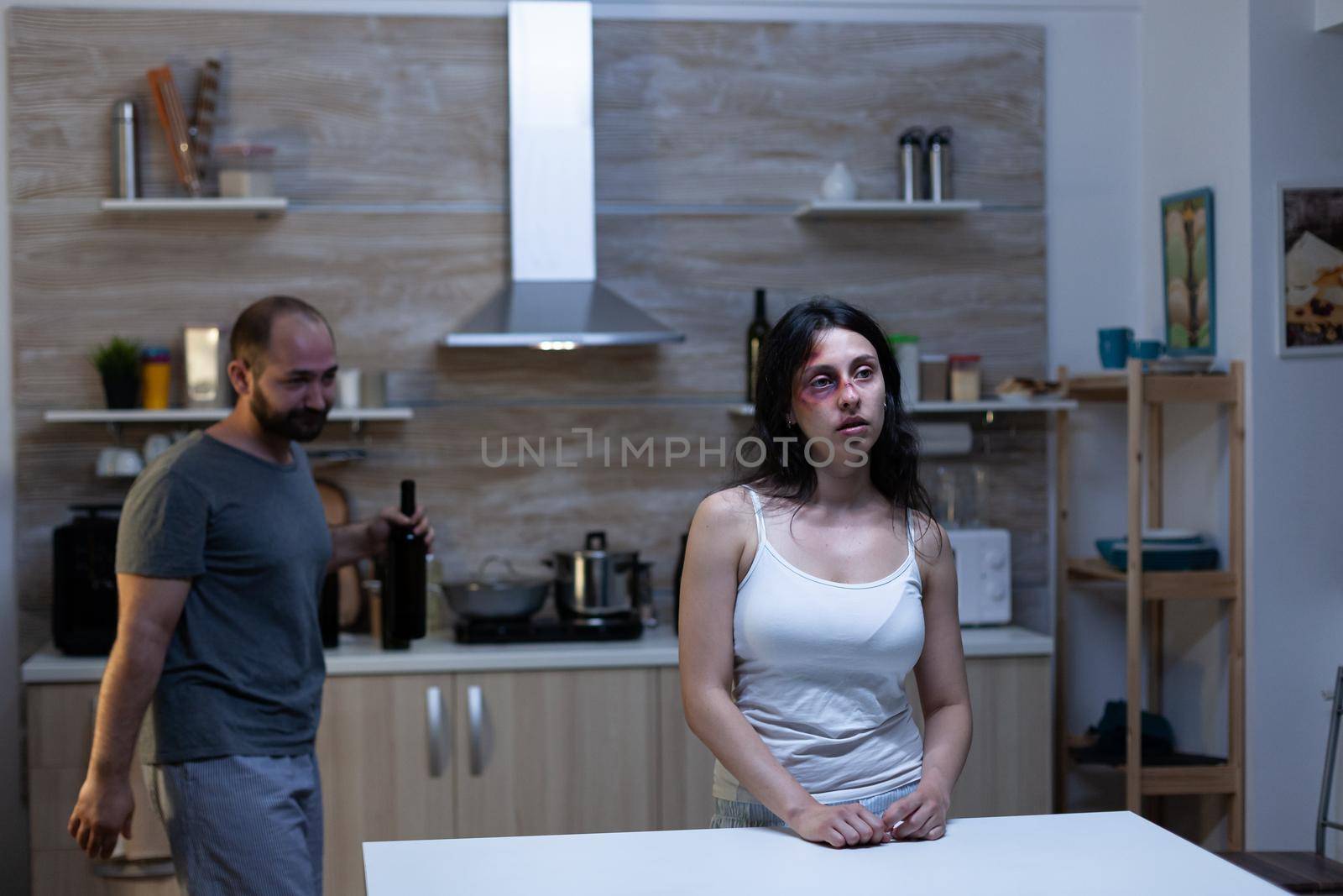 Abused woman sitting in kitchen with drunk husband dealing with aggression, conflict and alcohol addiction. Hurt bruised person having marriage problems with domestic violence