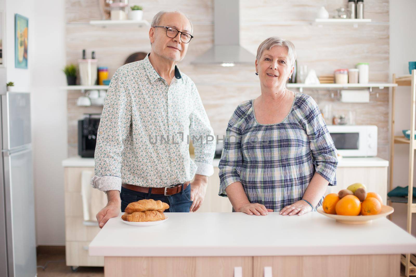 Portrait of old cheerful couple looking into camera in kitchen while preparing breakfast. Happy retired man and woman.