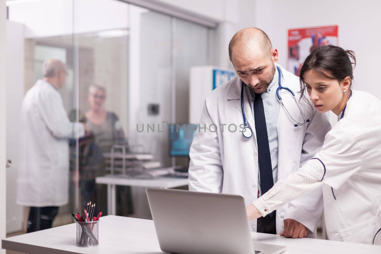 Team of young doctors working together in hospital checking patient evolution on laptop. Elderly aged woman and senior medic on clinic corridor. Physician wearing white coat and stethoscope.