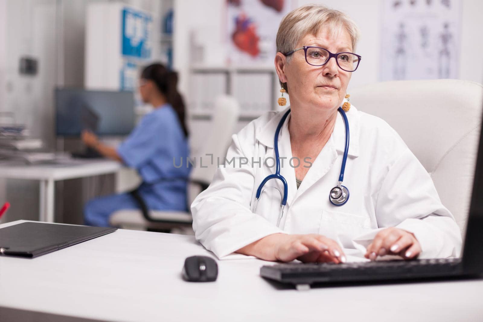 Portrait of experienced senior medic typing report on computer and nurse holding x-ray in the background.